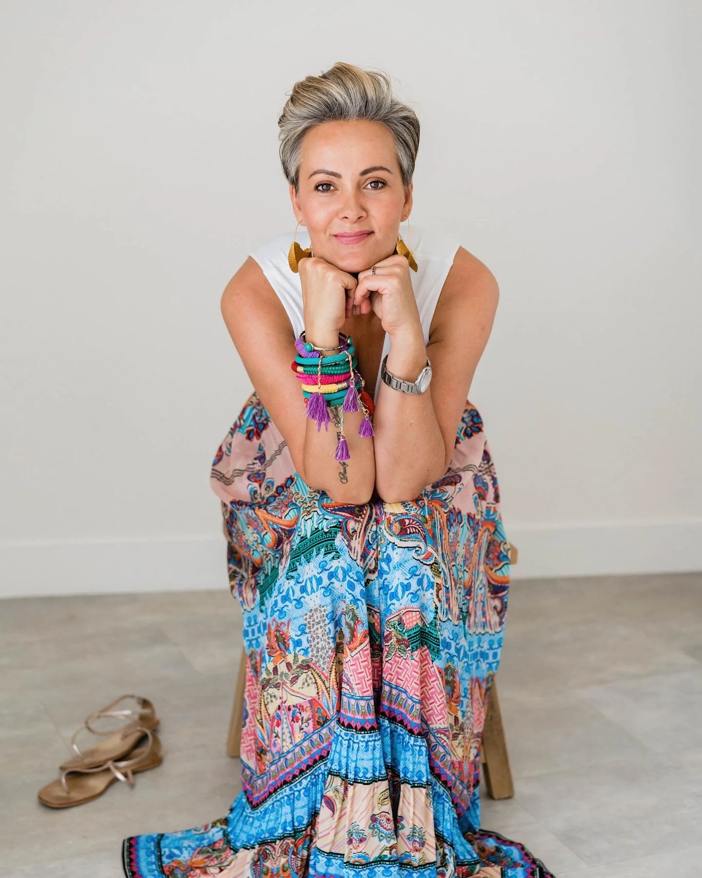 Nikki Gaskell: A woman with short gray hair sitting on a wooden stool, wearing colorful bangles, earrings, and a long, vibrant, patterned skirt with a white sleeveless top, looking at the camera with a relaxed pose.