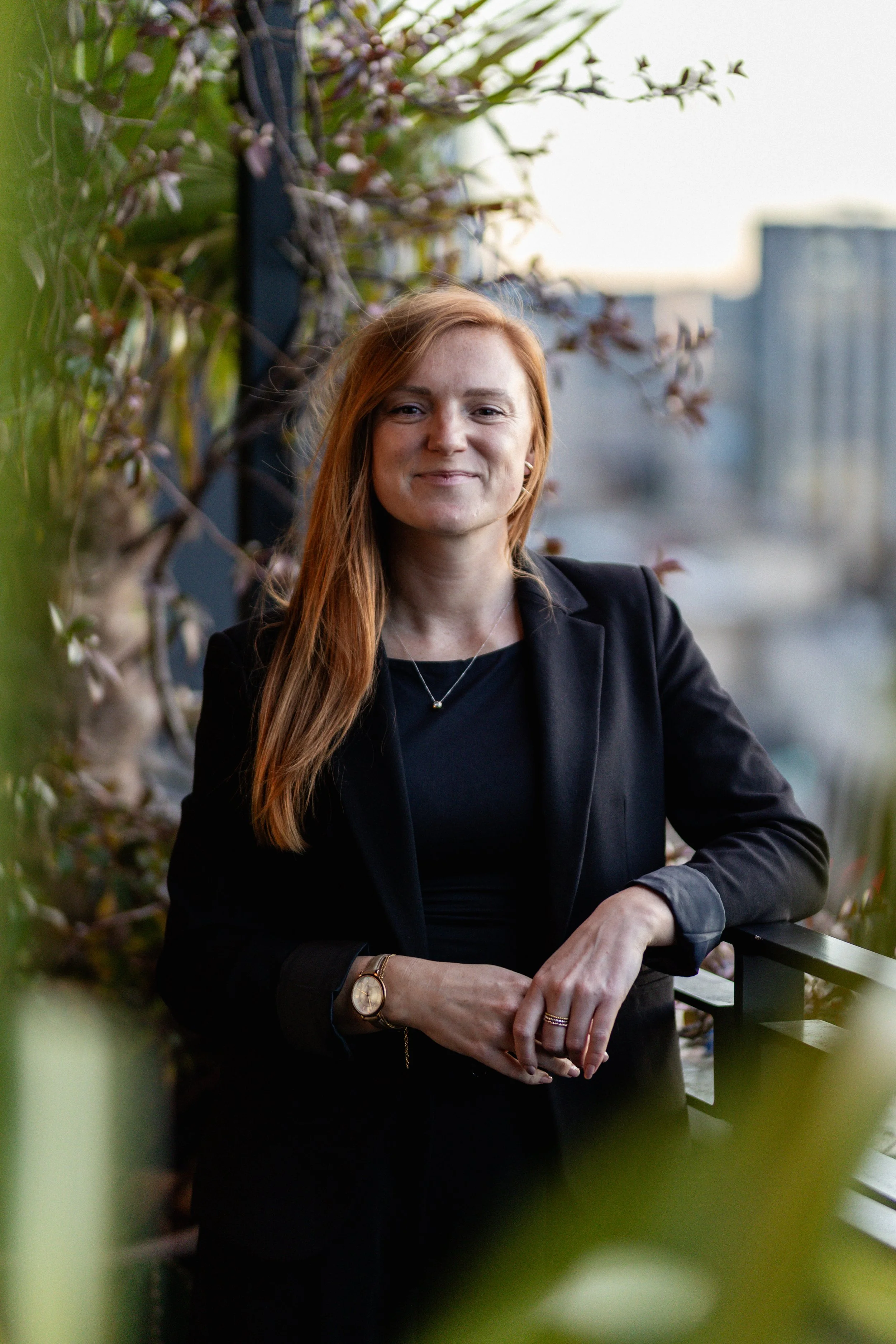 Erin Hegarty: A woman with long red hair, wearing a black blazer and black shirt, standing on a balcony with a city skyline in the background. She is smiling and has her hands resting on the balcony railing, surrounded by green foliage.