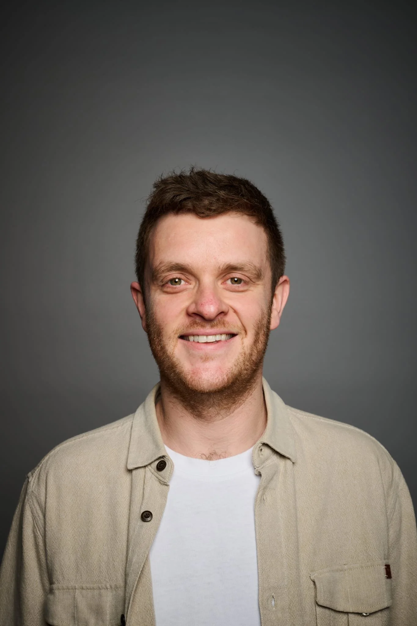 Nathan Cherokoff: Headshot of a young man with short brown hair and a beard, smiling, wearing a light-colored button-up shirt over a white T-shirt, against a dark gray background.