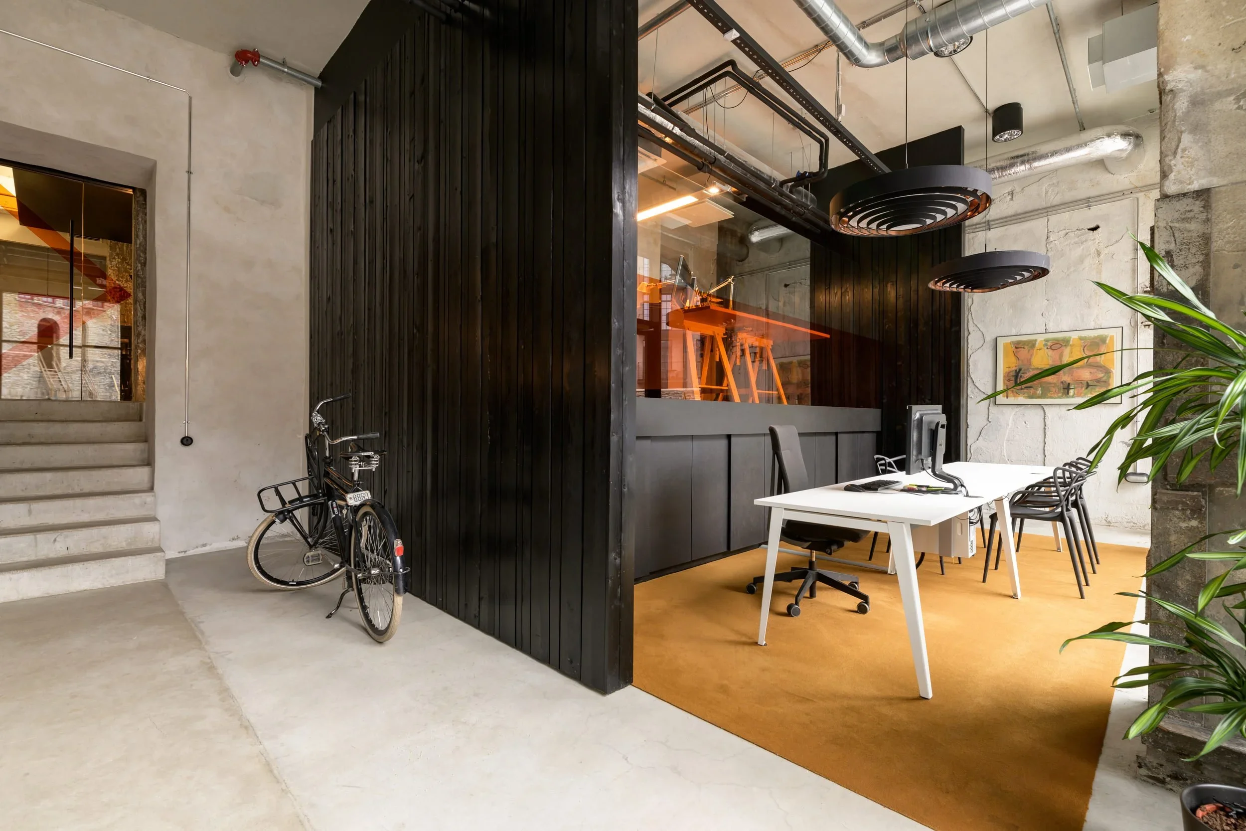 Office space with a white table, chairs, a black office chair, and a computer. There are two large black pendant lights, potted plant, artwork on the wall, and a black wooden partition with a glass window showing a room with desks and stools.