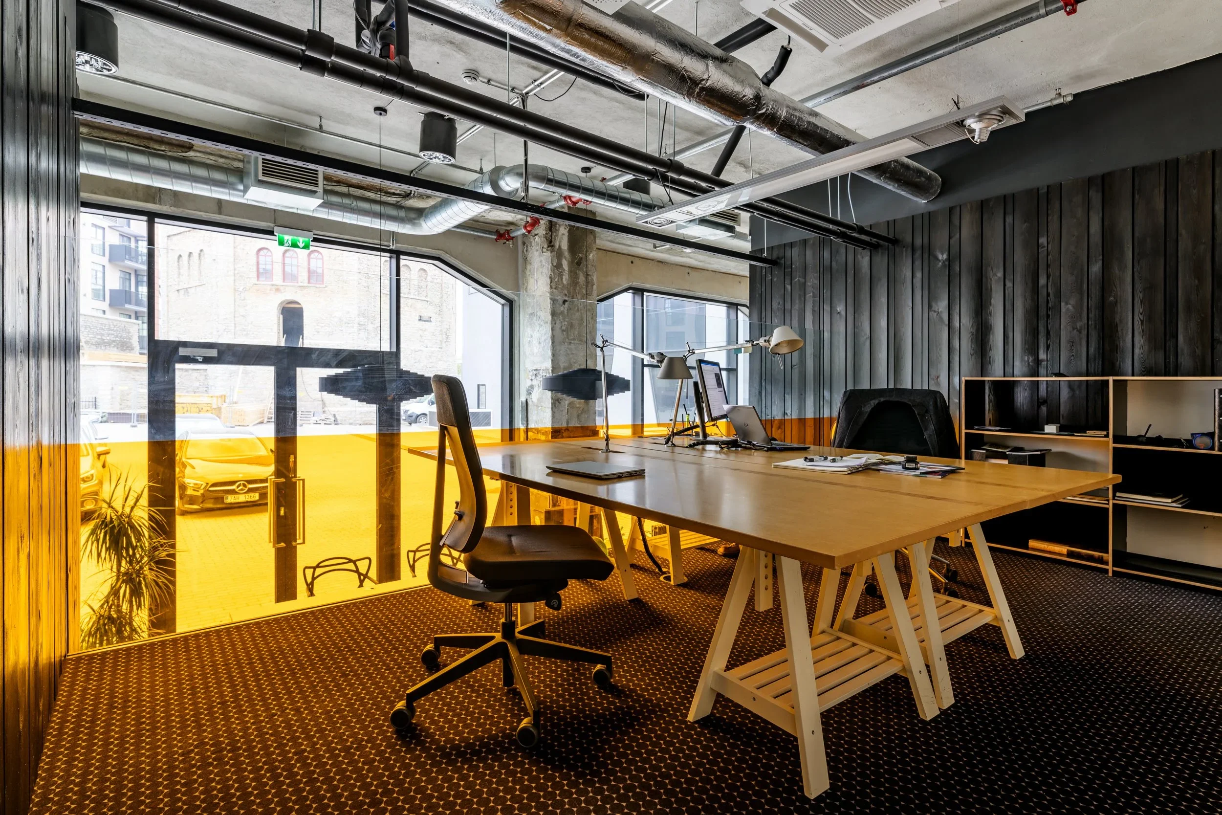 Modern office with a large wooden desk, rotating office chair, black wall panels, and floor-to-ceiling windows with a city view. Items on the desk include a laptop, desk lamps, and notebooks.