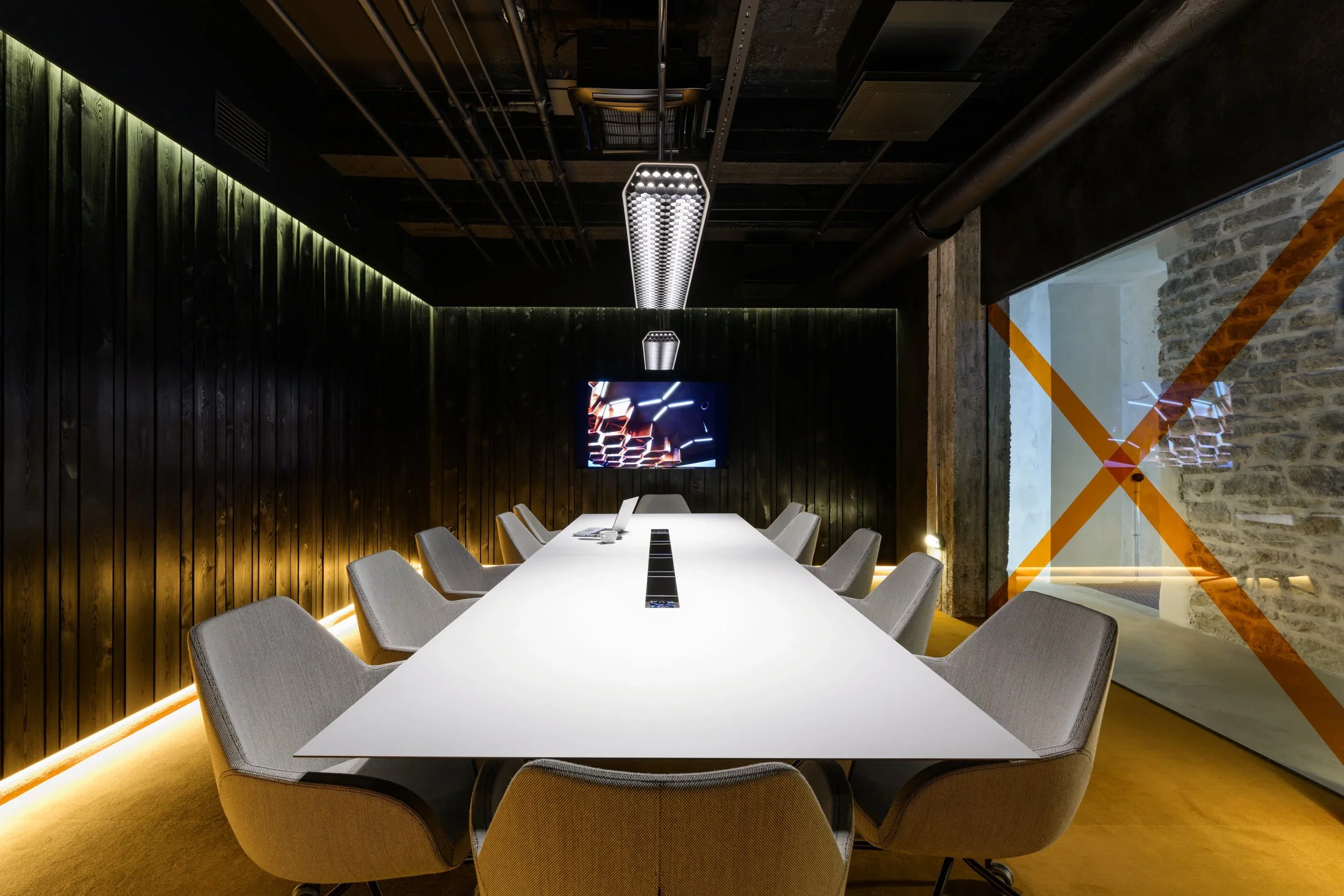 Modern conference room with a long white table, gray chairs, dark wood paneled walls, a wall-mounted TV, and contemporary lighting fixtures.