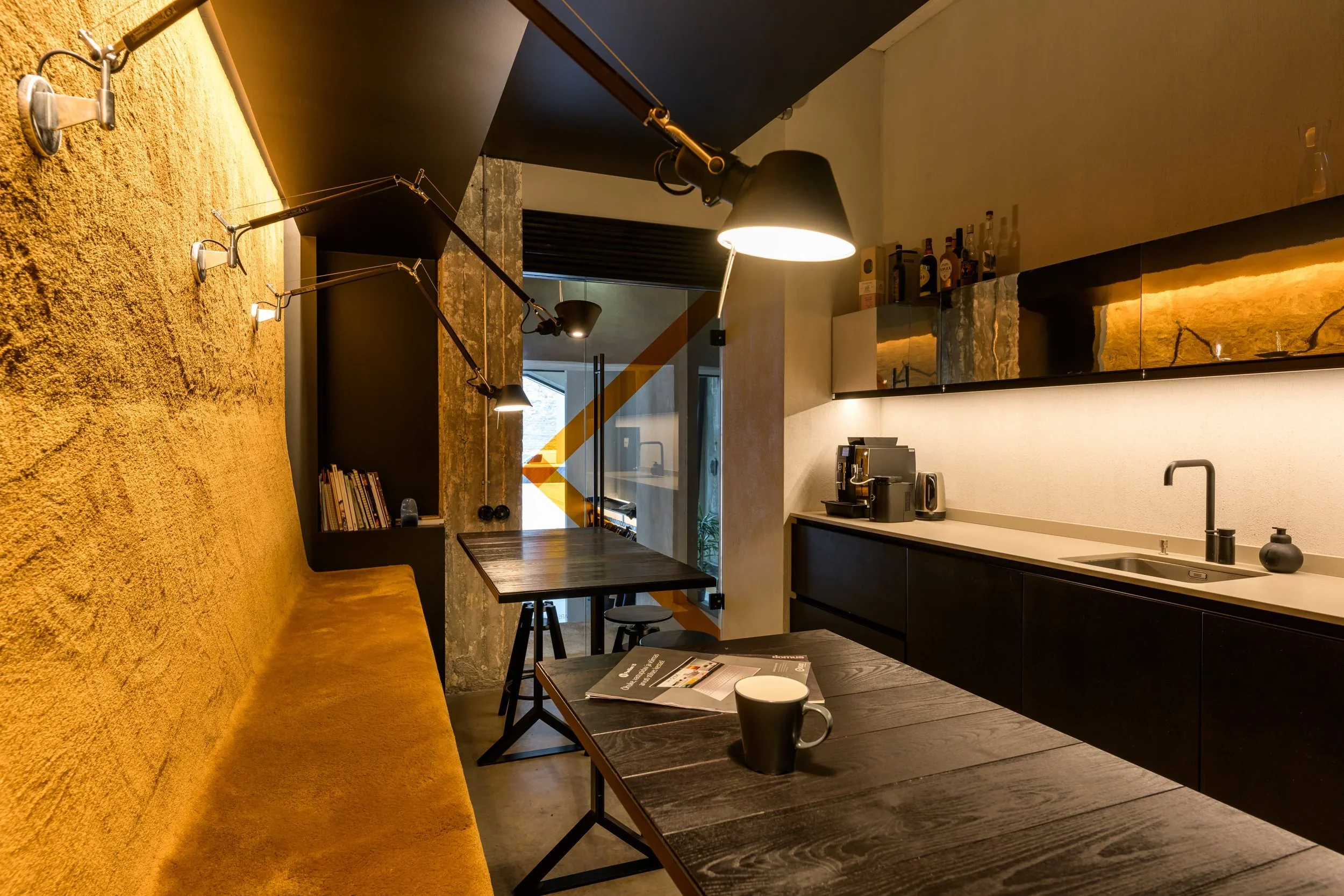 Cozy kitchen and dining area with black cabinets, yellow textured wall, black pendant and wall lamps, wooden table, bench, chair, coffee machine, bottles, and a mug.