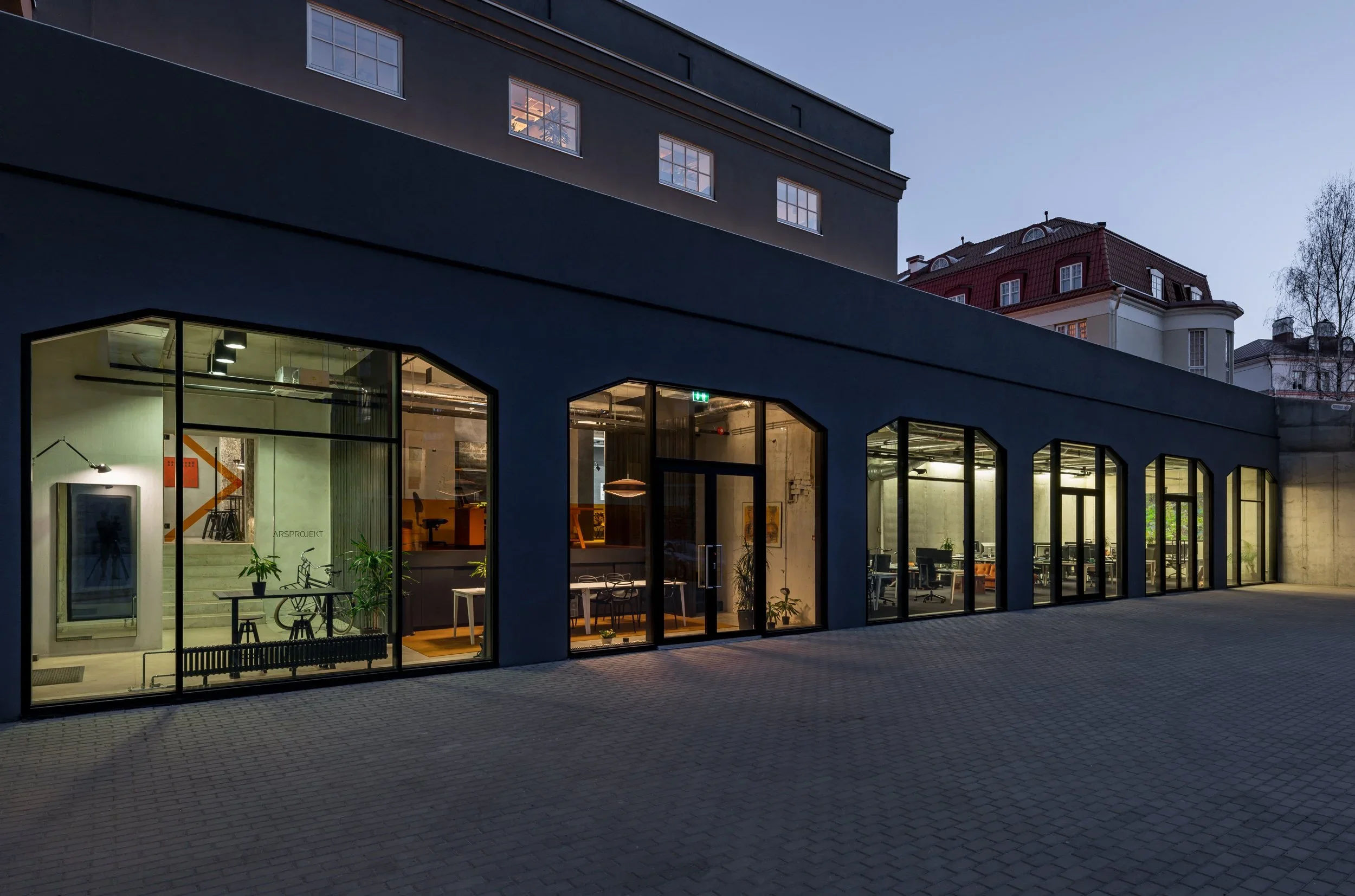 Modern office building with large glass windows illuminated from inside during dusk, with office furniture and plants visible.