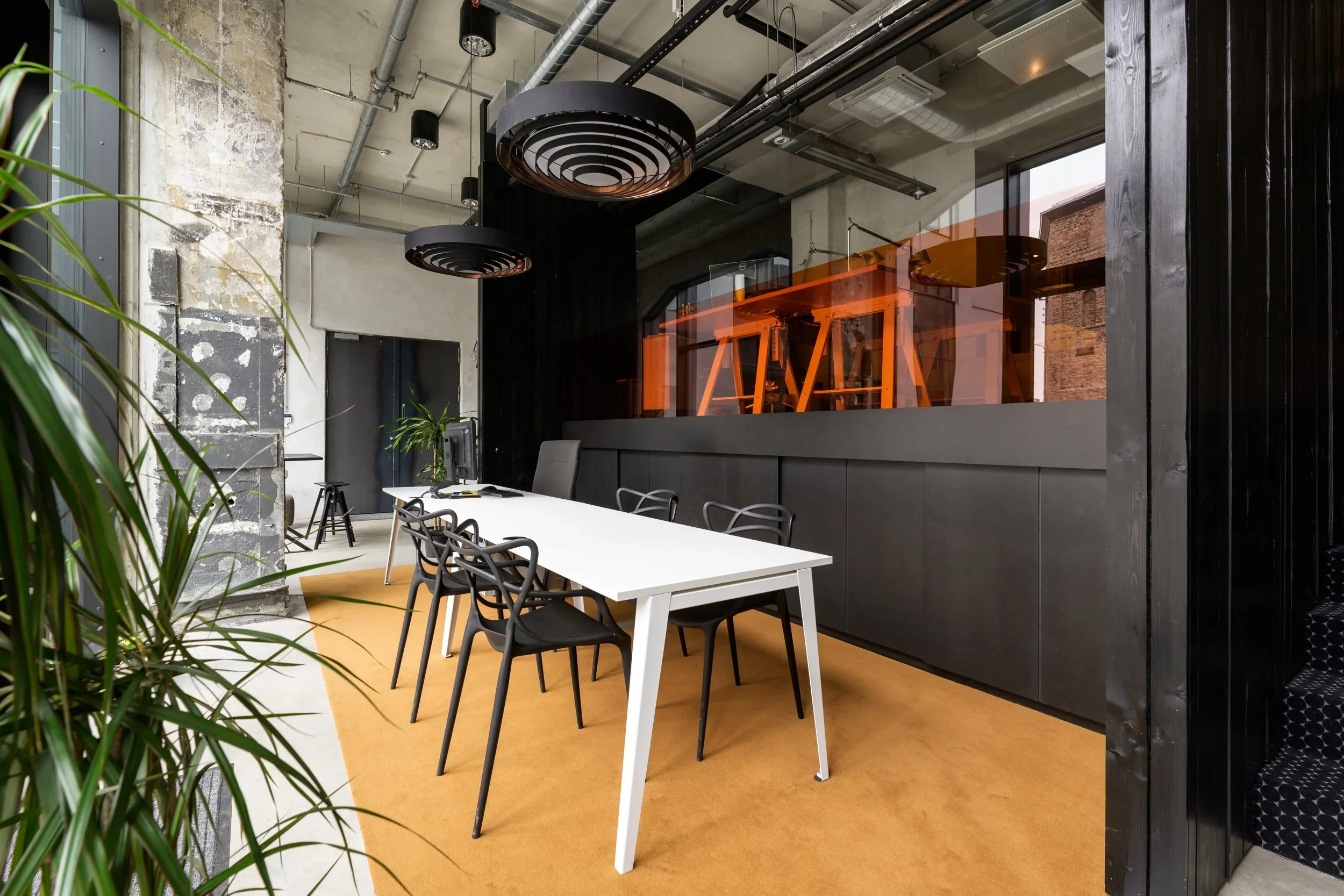 Modern office conference room with a white table, six black chairs, a computer monitor, potted plant, and industrial decor. There is an orange glass wall behind the table and black pendant lights hanging from the ceiling.