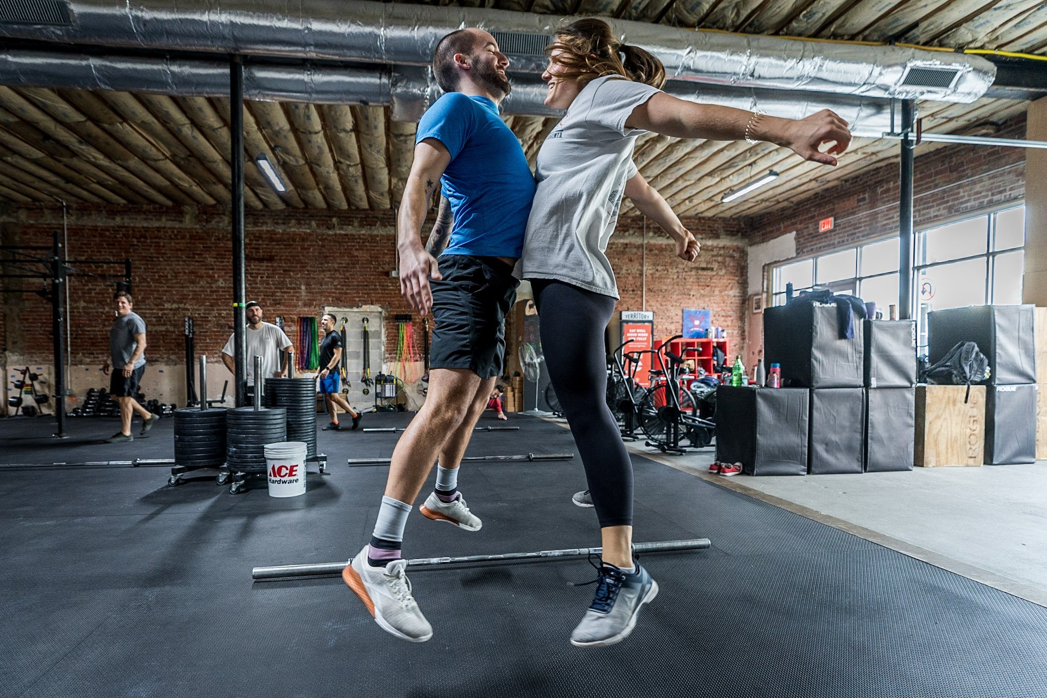 A man and a woman are jumping and hugging in a gym, with gym equipment and other people working out in the background.