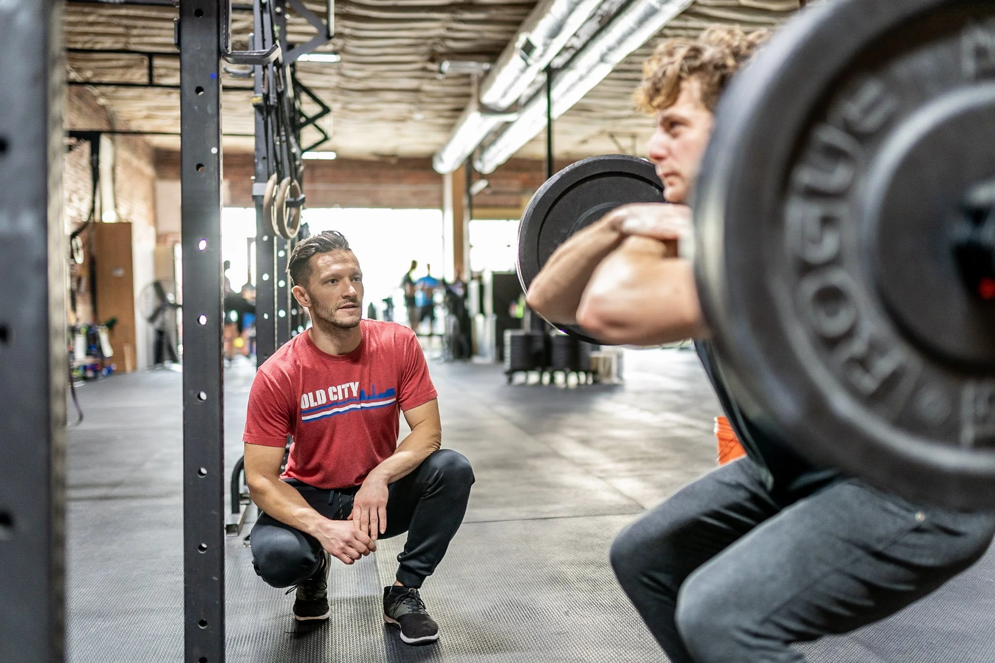 A man with curly hair squats while holding a barbell during a workout at a gym, with a trainer or coach kneeling and observing him.