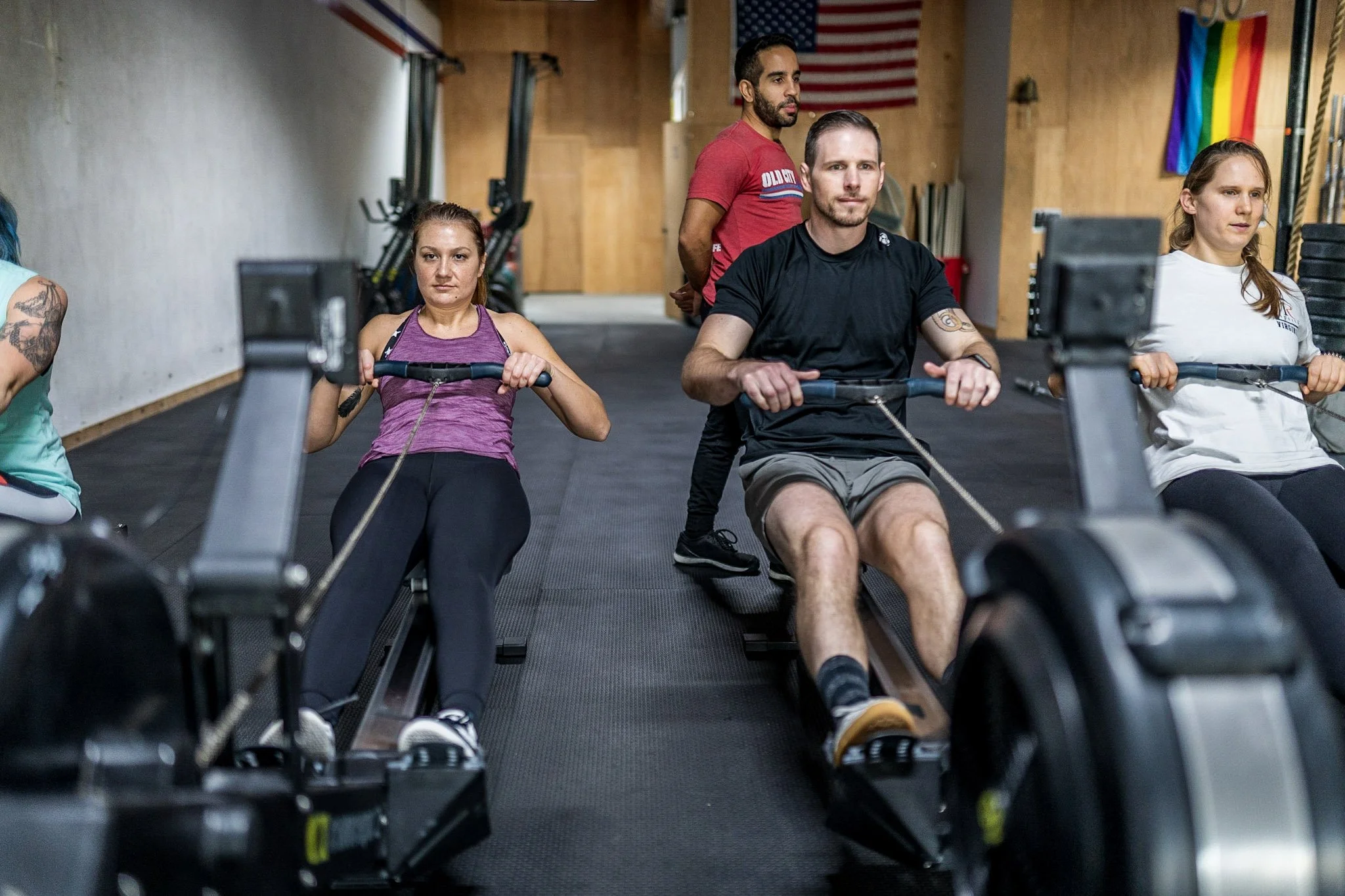 People working out on rowing machines in a gym, with a woman in a purple top on the left, a man in a black shirt and gray shorts in the middle, and a woman in a white shirt on the right. There are black mats on the floor and American and rainbow prid
