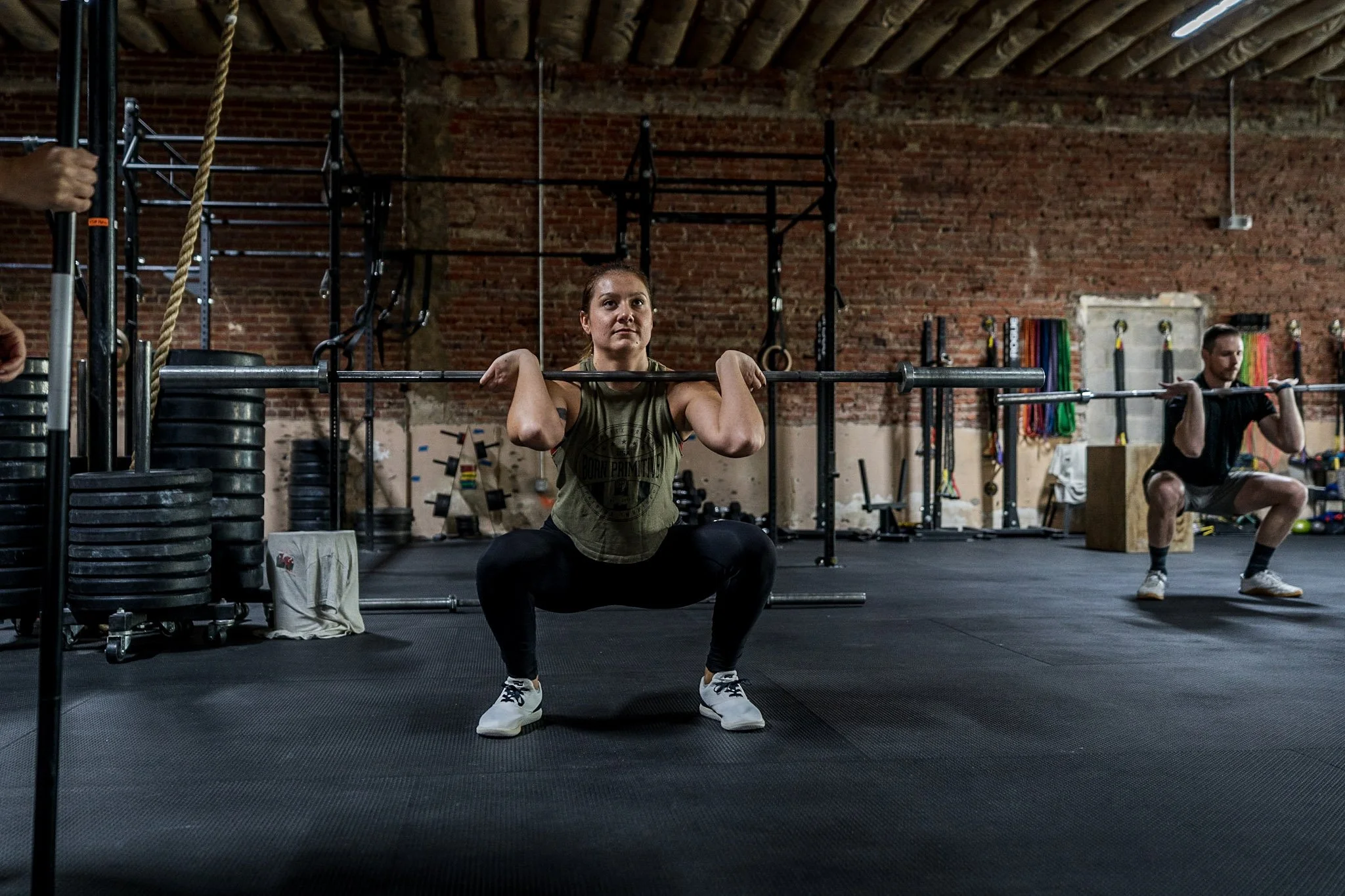 A woman performing a squatting exercise with a barbell at a gym, with a man in the background also lifting a barbell.
