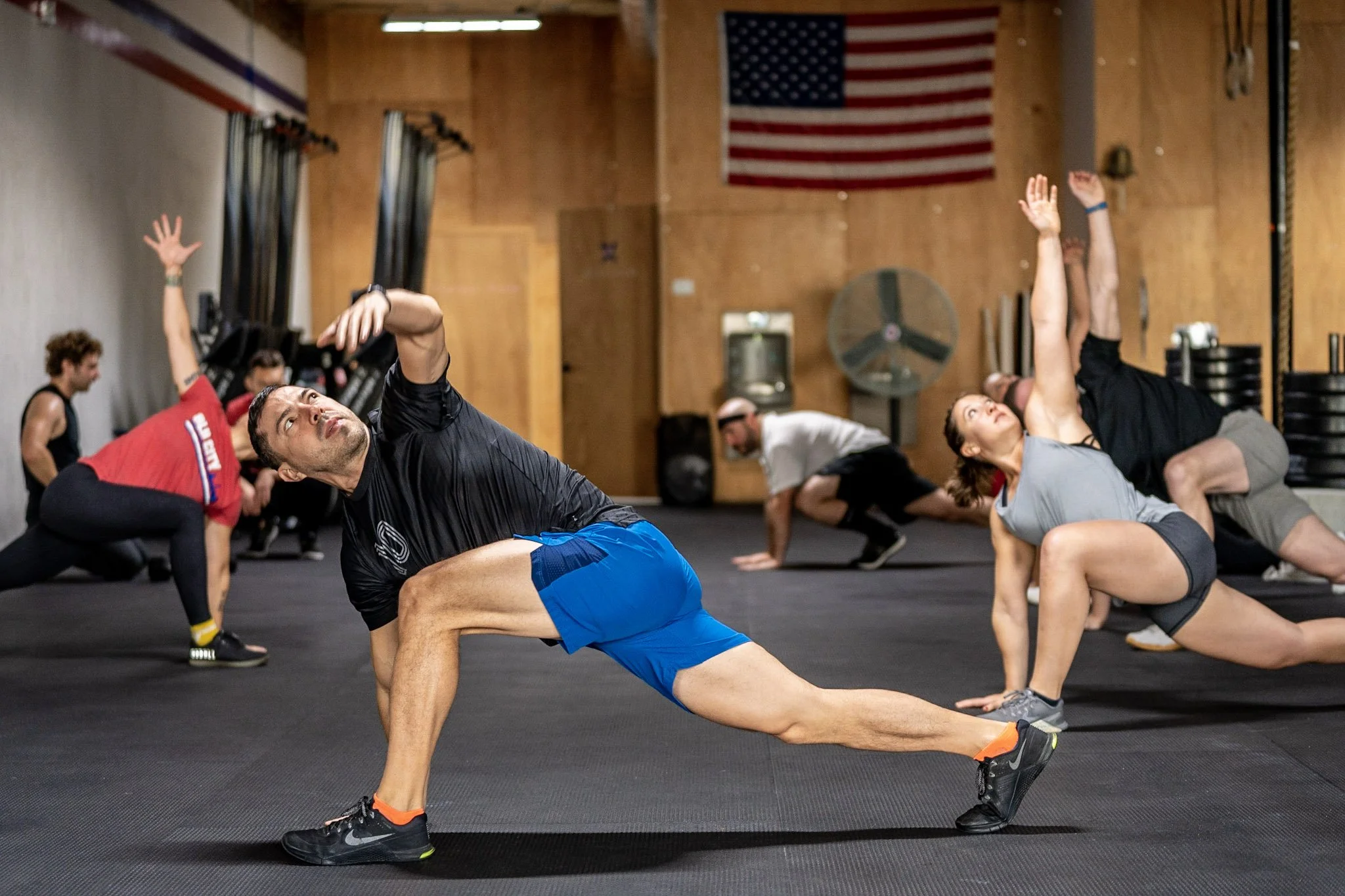 A group of people in athletic clothing doing various stretches or yoga poses in a gym with wood walls and an American flag hanging on the wall.