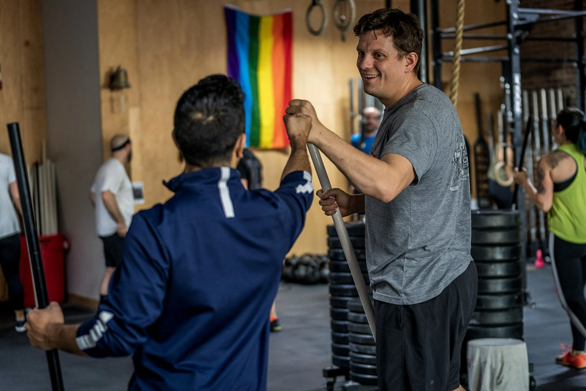 Two men shaking hands inside a gym, one holding a barbell, with a rainbow flag on the wall in the background and other people exercising.