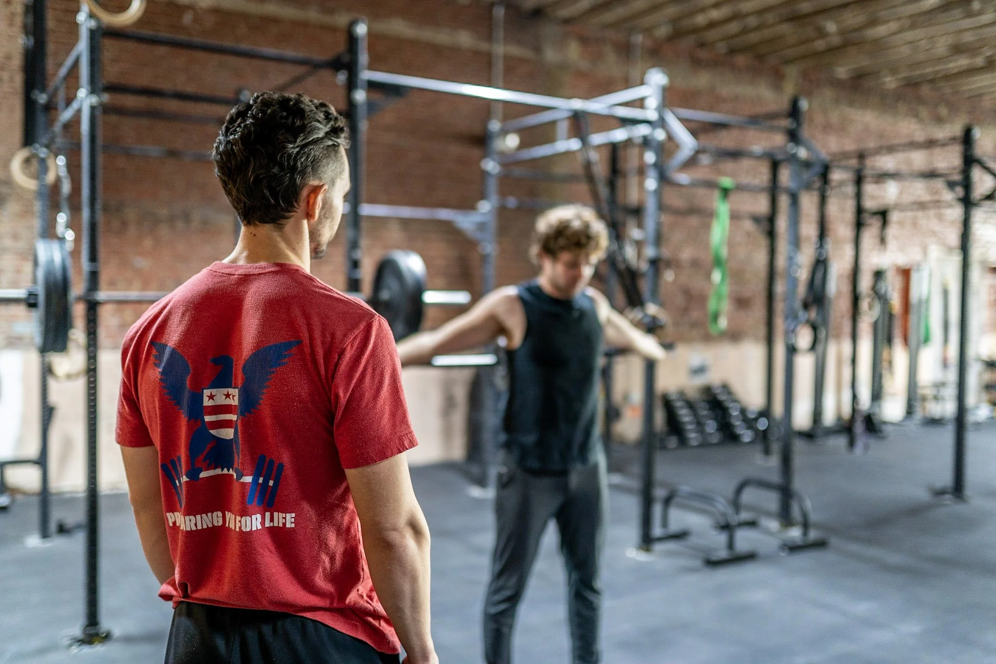 Two men in a gym with one man standing in the foreground wearing a red T-shirt and the other man in the background doing a shoulder press exercise with weights.