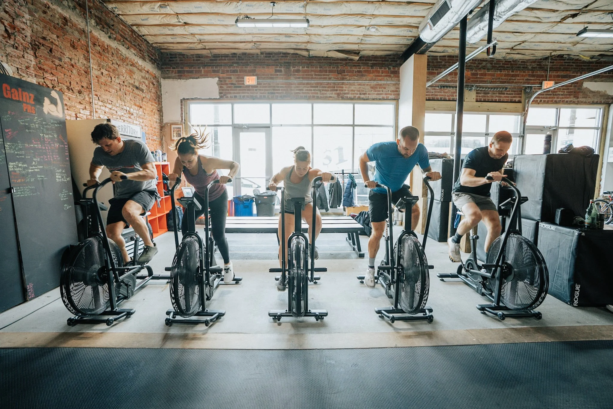 Group of five people working out on air bikes in a gym with exposed brick walls and large windows.