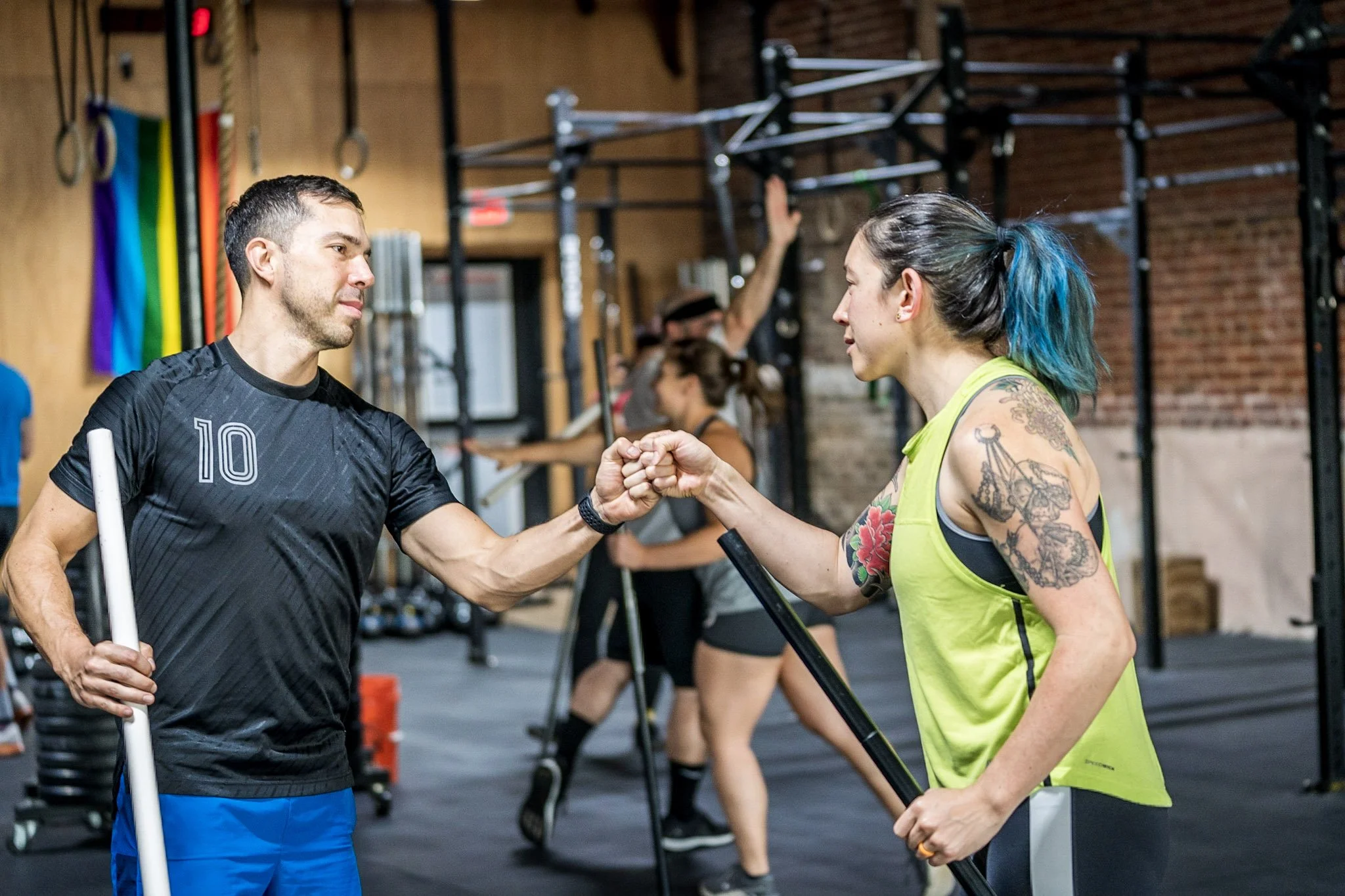 A man and woman in a gym fist bump each other, with several other people exercising in the background.