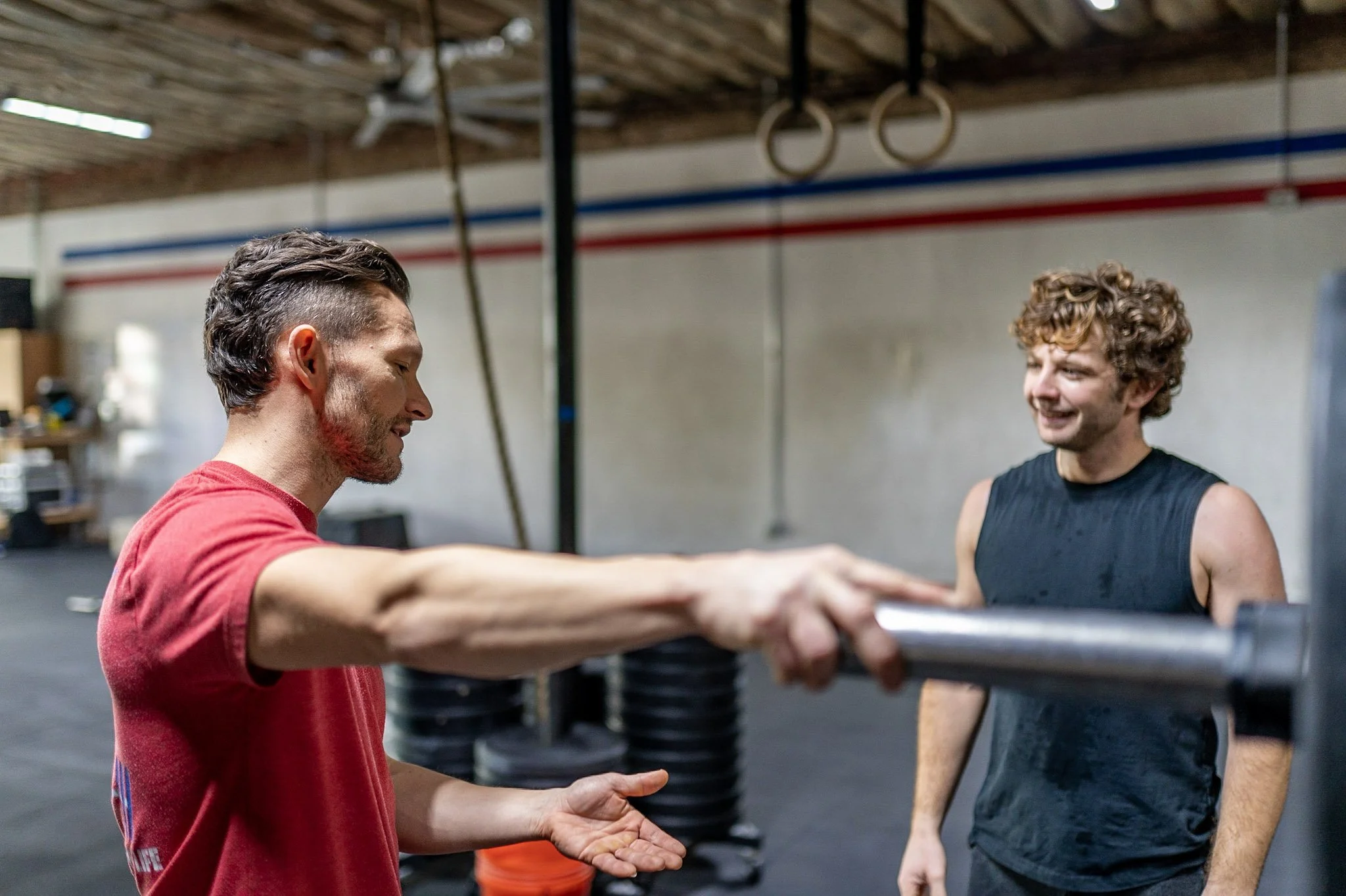 Two men at a gym. One man in a red shirt is holding a barbell, assisting or instructing the other man in a black workout tank top, who is smiling.
