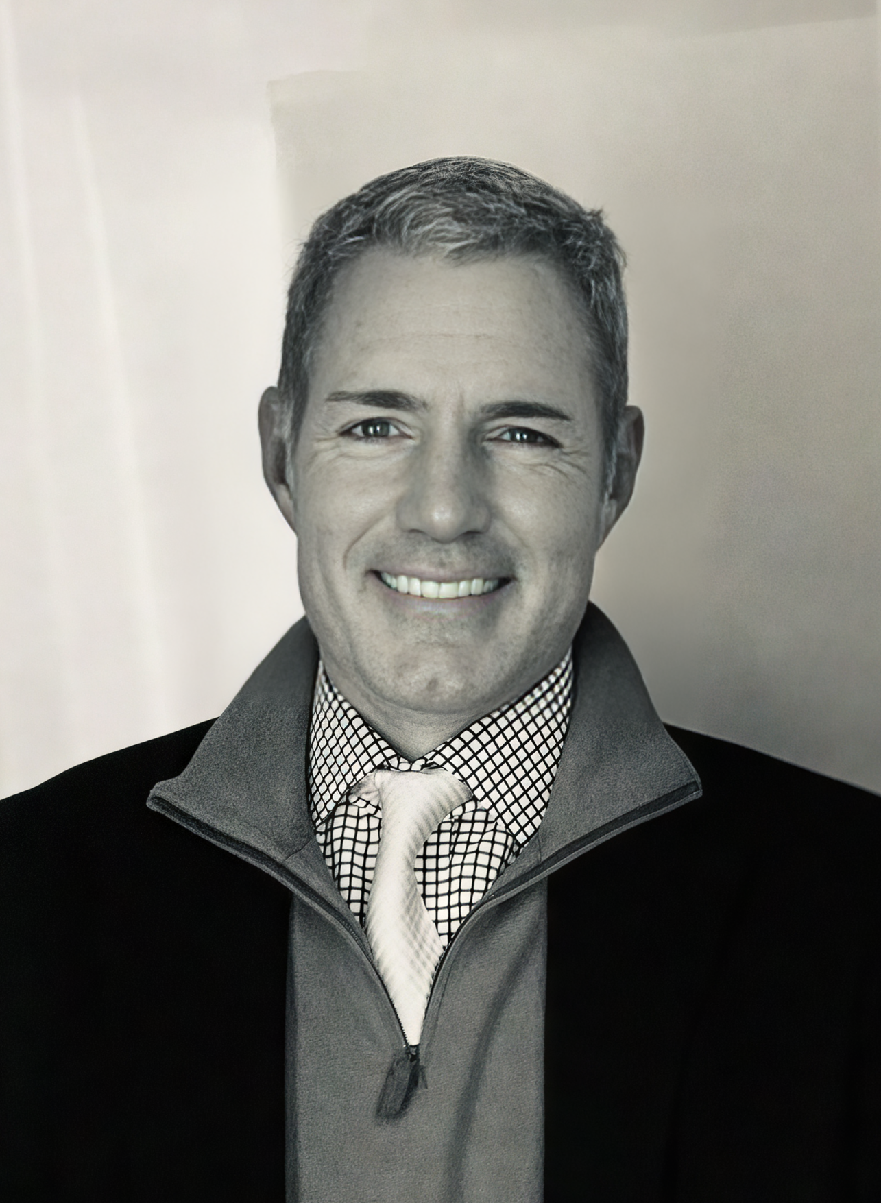 Black and white headshot of smiling man in dress shirt, tie, and zip-up sweater against plain background.