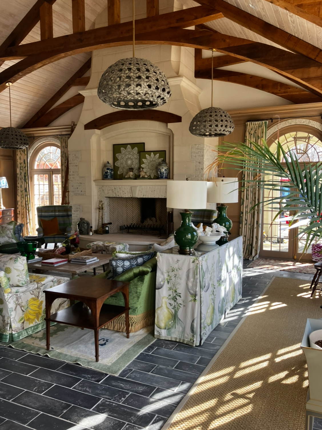 A cozy living room with a high, vaulted wooden ceiling and exposed beams. There are three pendant lights hanging from the ceiling, a white stone fireplace with decorative objects on the mantel, and large arched windows with floral curtains letting in