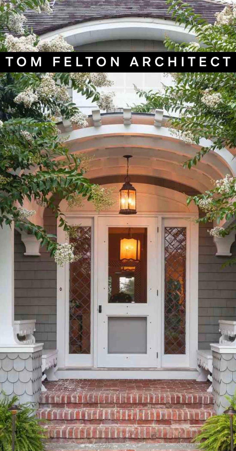 Front porch of a house with brick steps, a white door with glass panels, surrounded by greenery and flowering plants, with a hanging lantern light above the door.