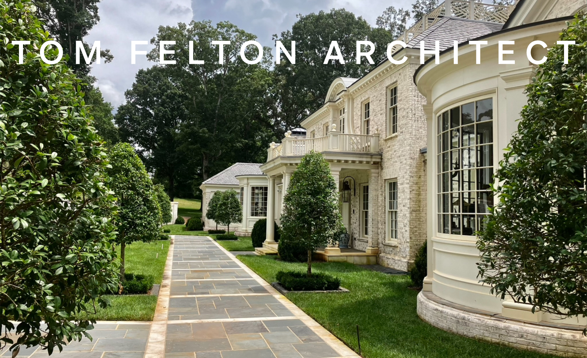 Front yard of a large, elegant house with a stone and cream exterior, large bay window, trees, and a paved walkway.