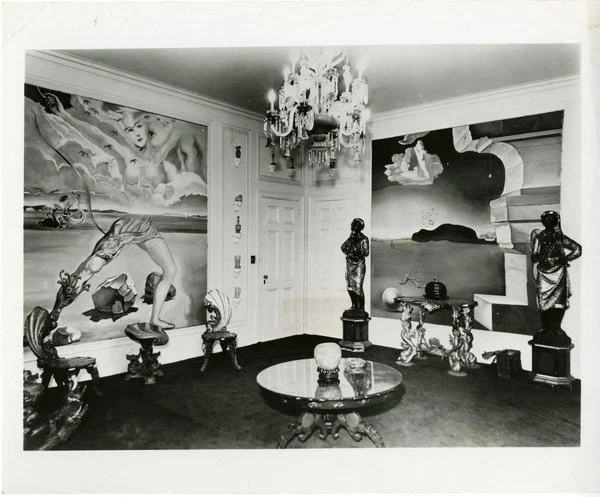 A black-and-white photo of an art-filled living room with ornate furniture, a chandelier, and large paintings on the walls.