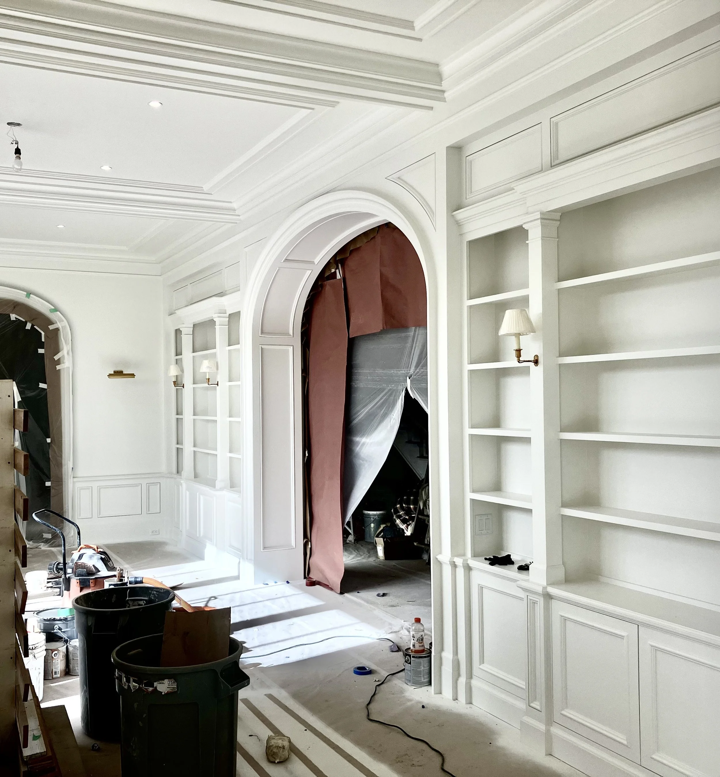 Interior of a house under renovation, showing a white built-in bookshelf and a partially covered arched doorway leading to another room. Construction tools and supplies are scattered on the floor.