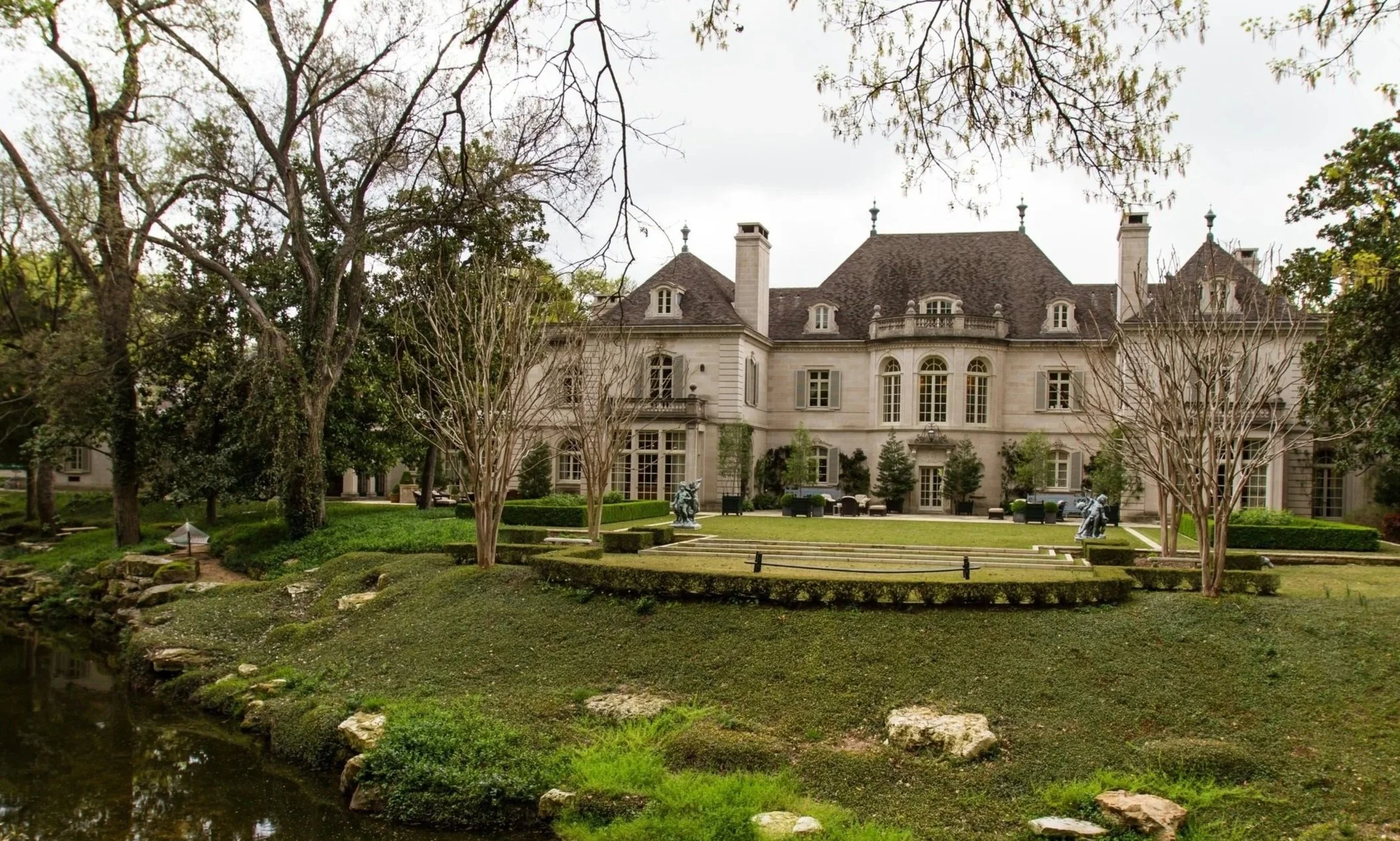 Large mansion with multiple chimneys and arched windows, surrounded by manicured lawns, trees, and sculptures, with a body of water in the foreground.