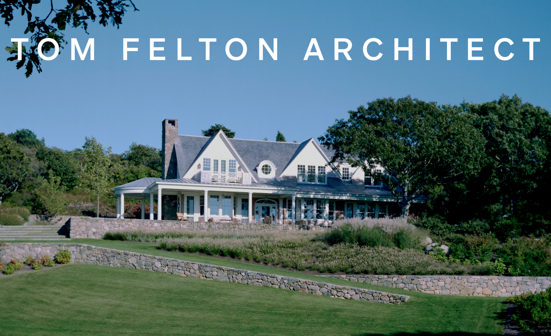A large, white, Victorian-style house on a hill with a green lawn, stone retaining walls, and trees, under a blue sky.