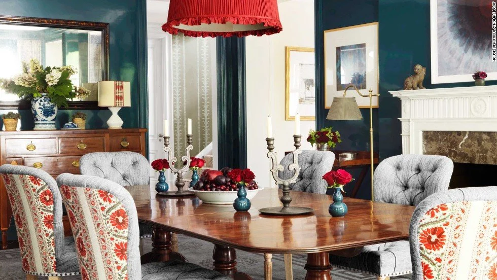 A dining room featuring a wooden table with five gray upholstered chairs, red roses in blue vases, two candelabras, and a bowl of red and purple fruit. There is a wooden sideboard with a flower arrangement, a mirror, and a lamp. The room has dark blu
