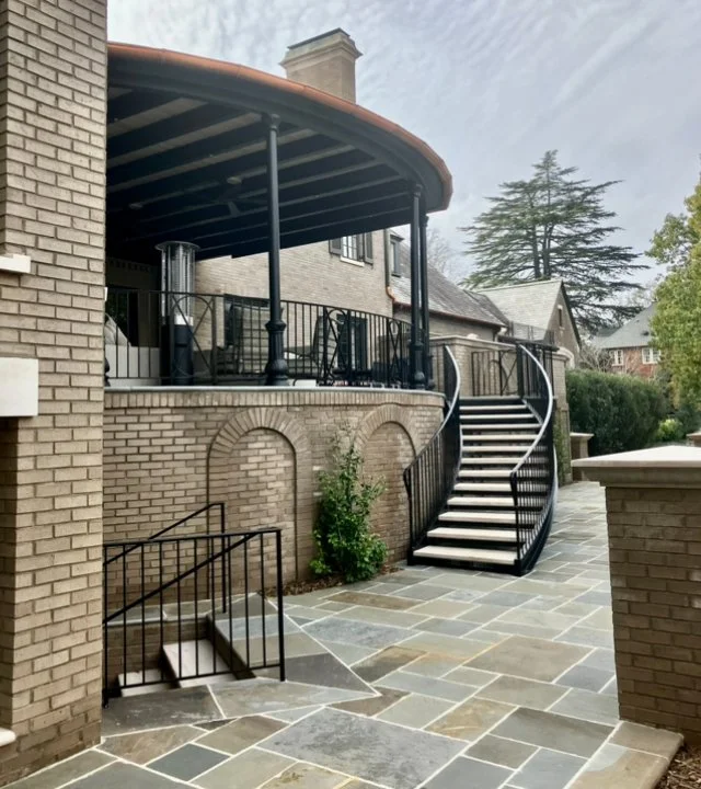 Curved staircase leading up to an elevated patio with black railing, brick house with arched brick details, and a landscaped yard with trees.