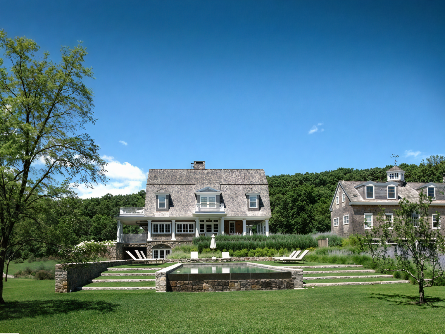 A large house with a sloped roof and enclosed porch with white railings, situated on a lush green lawn with trees and a clear blue sky in the background.