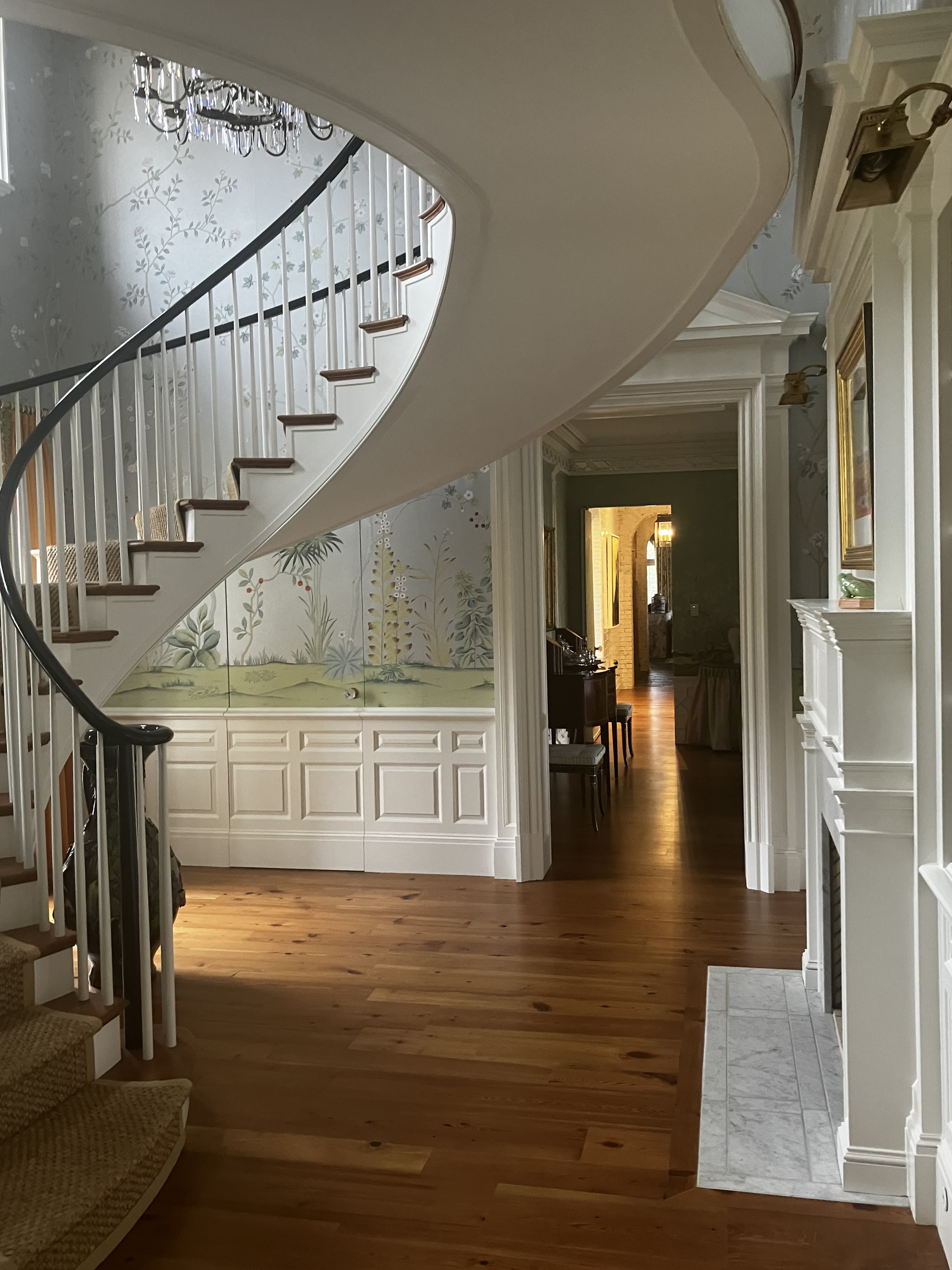 Interior view of a house with a curved staircase, wooden flooring, floral and wainscoting wall decor, and a hallway leading to another room with warm lighting.