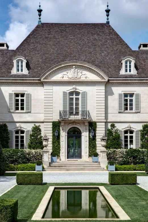 Front view of an elegant, historic mansion with a steep roof, dormer windows, ornate architectural details, and a well-maintained front garden with a water feature.