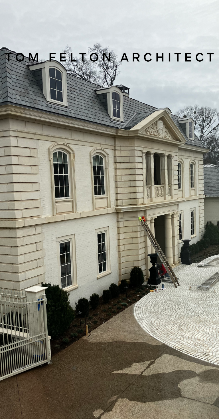 View of a large, elegant, white three-story house with arched windows, decorative pediments, and dormer windows on a gray, cloudy day. There is a ladder leaning against the house and construction tools nearby, with some landscaping and a circular pav