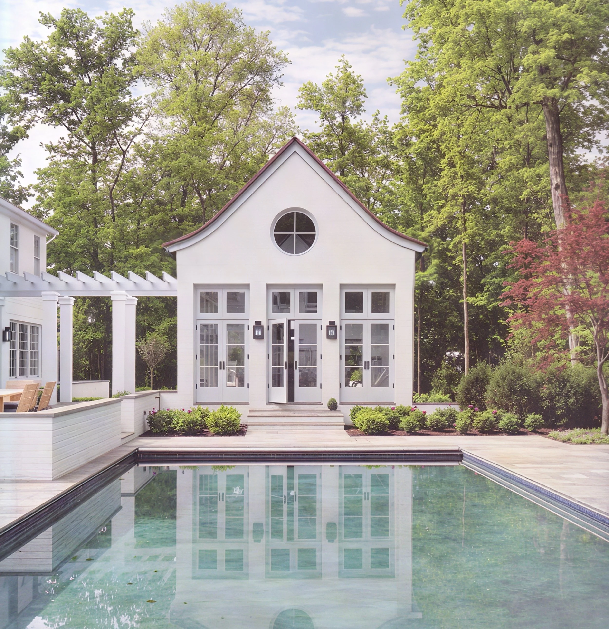 White modern pool house with glass double doors, circular window, and a pool in the backyard, surrounded by trees and garden greenery.