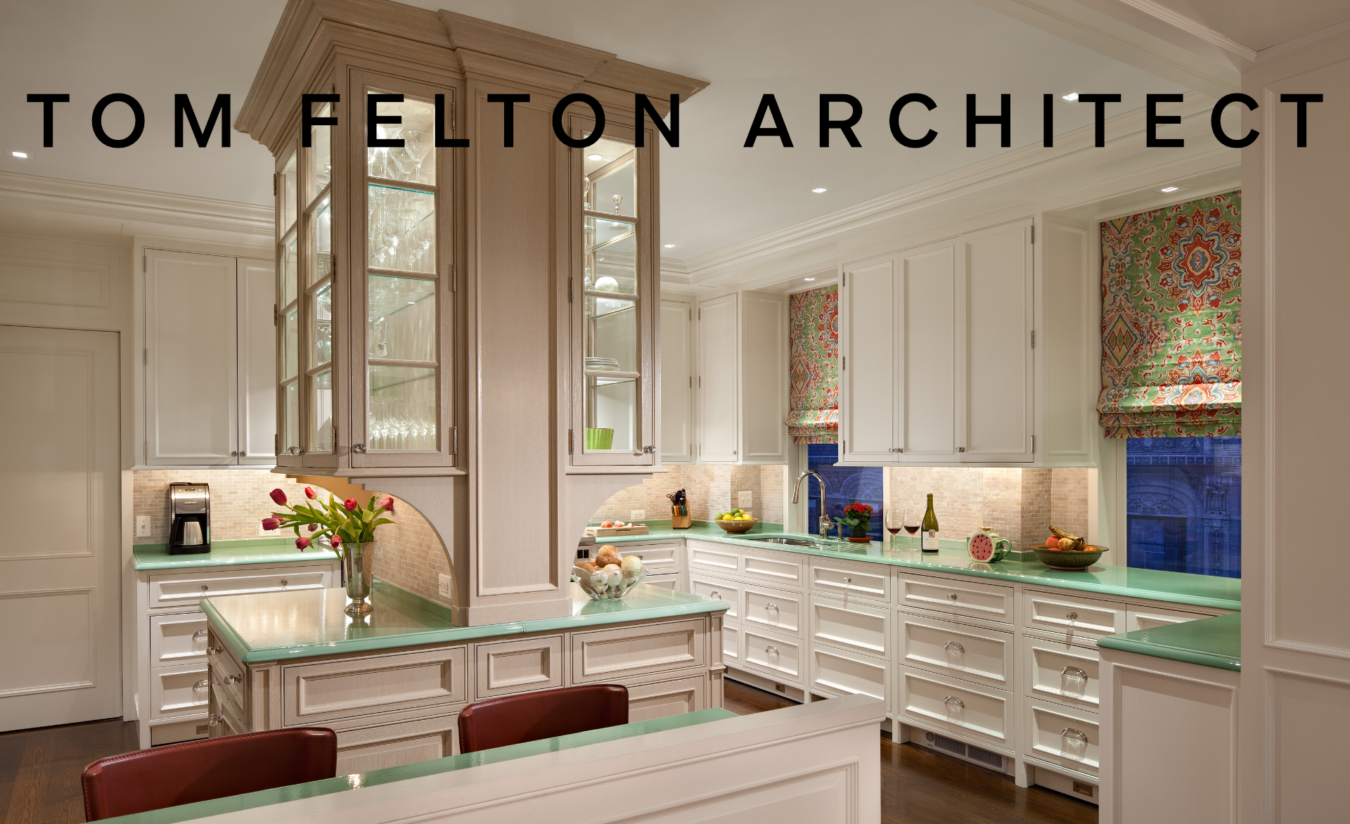 Interior view of a kitchen with white cabinetry and green countertops. There are decorative window coverings with colorful patterns and various kitchen items, including a bowl of fruit, a bottle of wine, and a vase of flowers.
