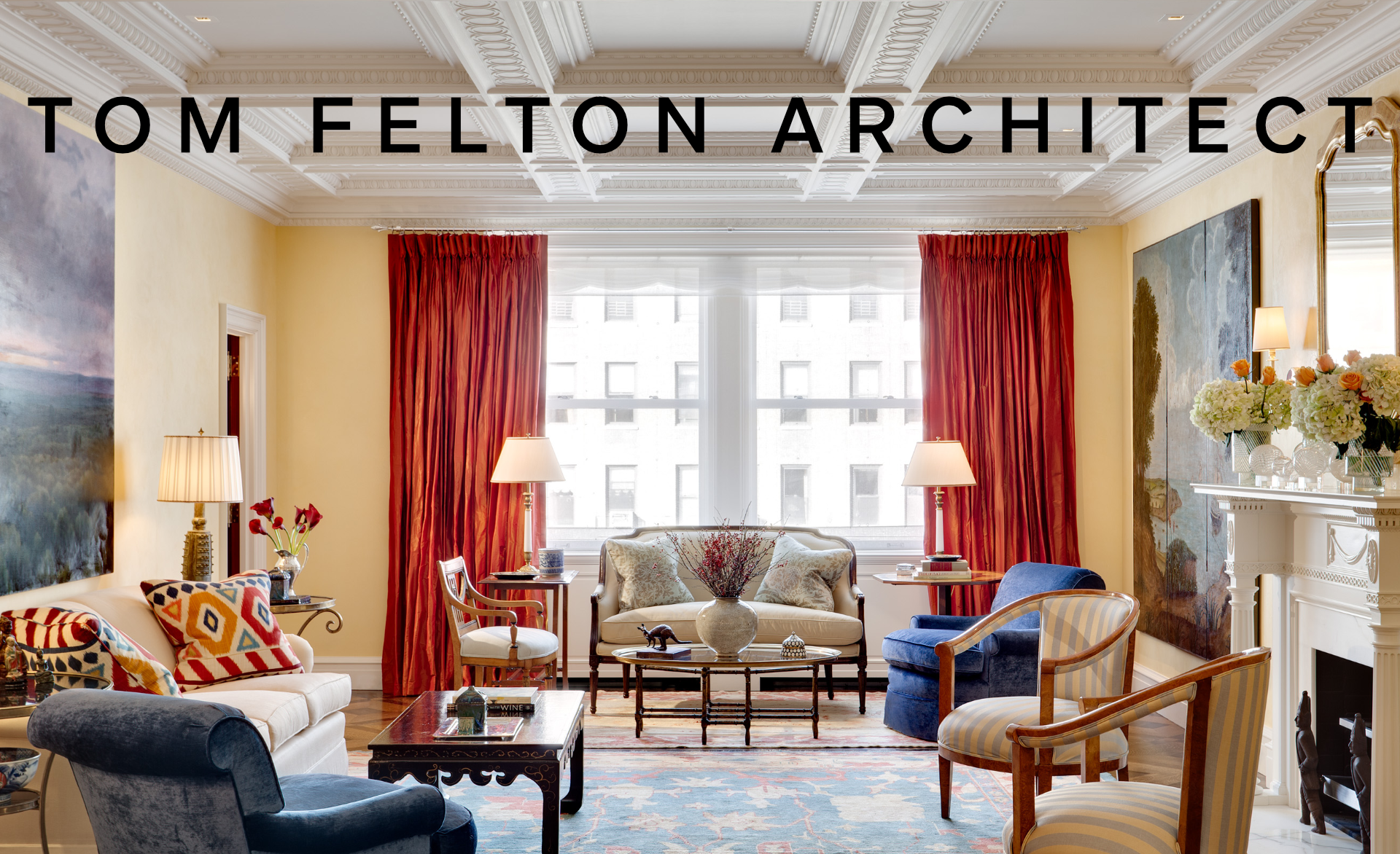 Elegant living room with cream walls, red curtains, a chandelier ceiling, and classic furniture including beige and blue armchairs, a white sofa with colorful pillows, and a fireplace decorated with flowers and vases. The room has large windows allow