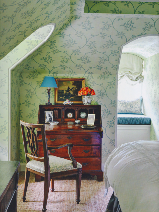 Vintage bedroom corner with green floral wallpaper, wooden desk with decorative items, framed photos, and a blue-shaded lamp. A cushioned window seat is visible in a nook, with part of a bed in the foreground.