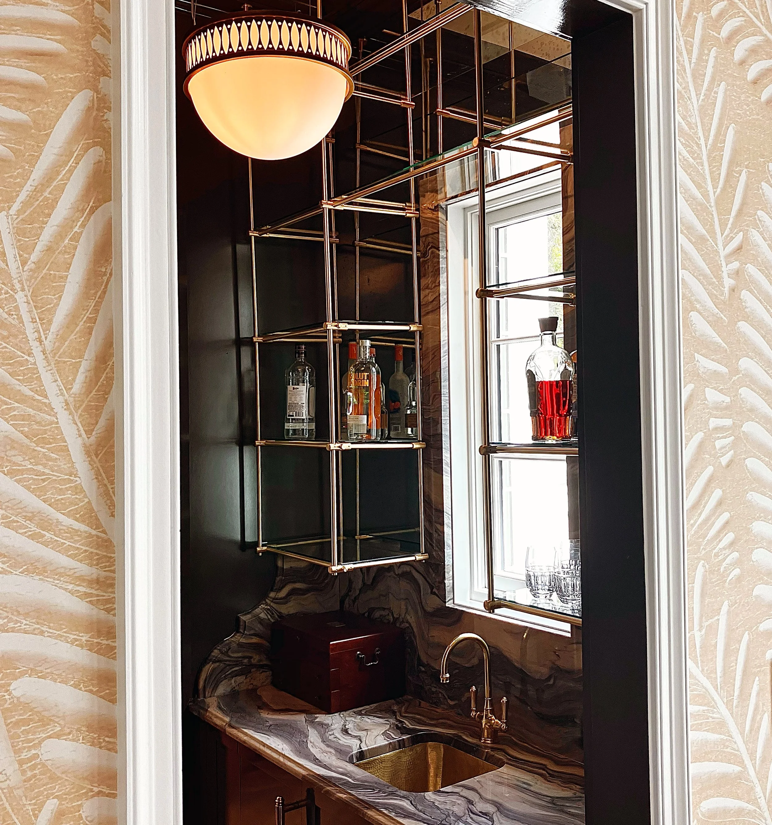 A small bar area with a marble countertop, a bronze sink faucet, and a window. There are shelves with bottles of liquor, illuminated by a ceiling light. The surrounding walls have beige patterned wallpaper.