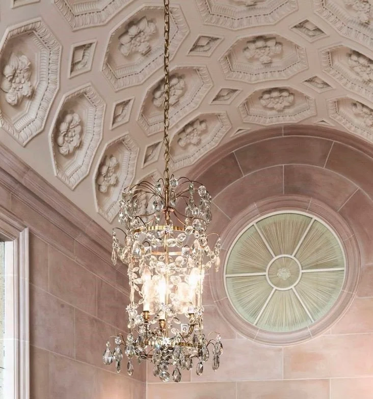 An ornate ceiling with floral and geometric patterns, a large circular decorative window, and a hanging crystal chandelier in a building interior.