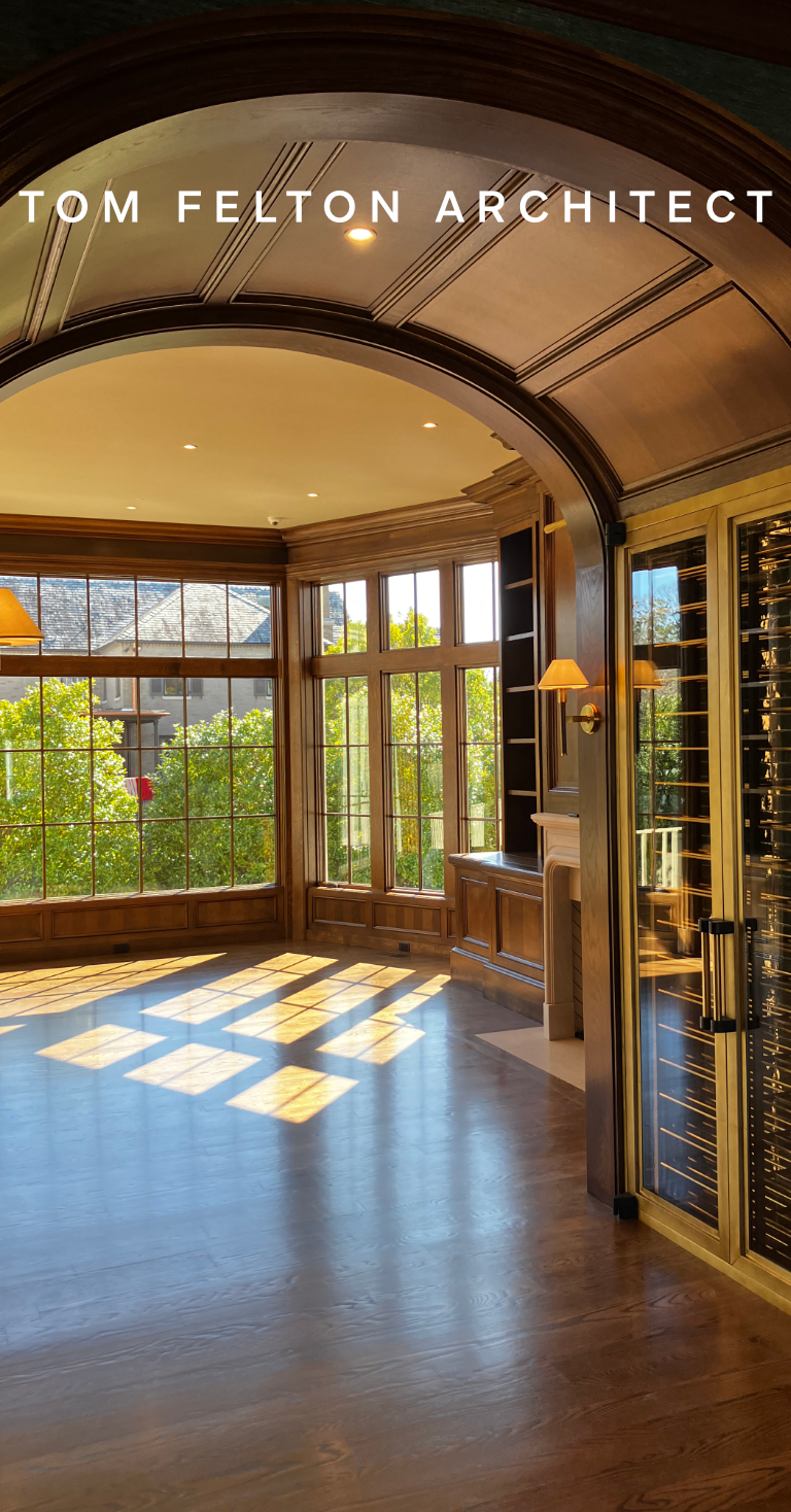 Sunlight streams through large windows into an elegant wooden interior space with hardwood floors, built-in shelving, and a gold and glass wine cooler, designed by architect Tom Felton.