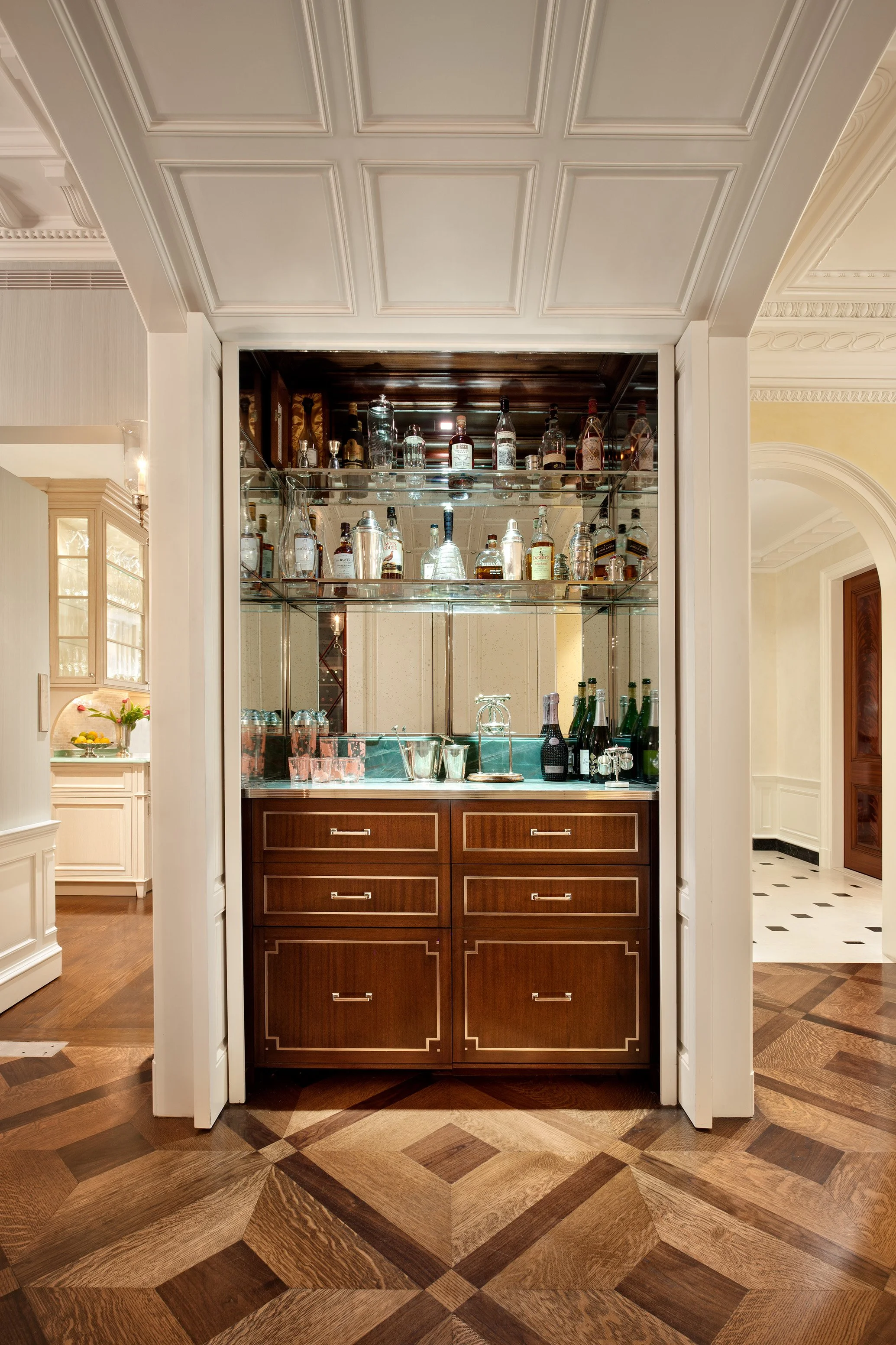 A built-in bar with glass shelves displaying various bottles of alcohol, situated in a room with ornate white ceiling and wood-paneled floor.