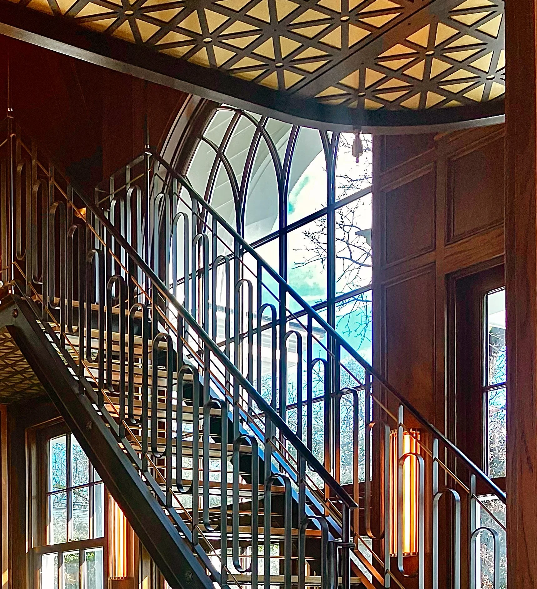 Interior view of a staircase with black metal railing, large arched windows, and wood-paneled walls. The ceiling has a decorative geometric pattern.