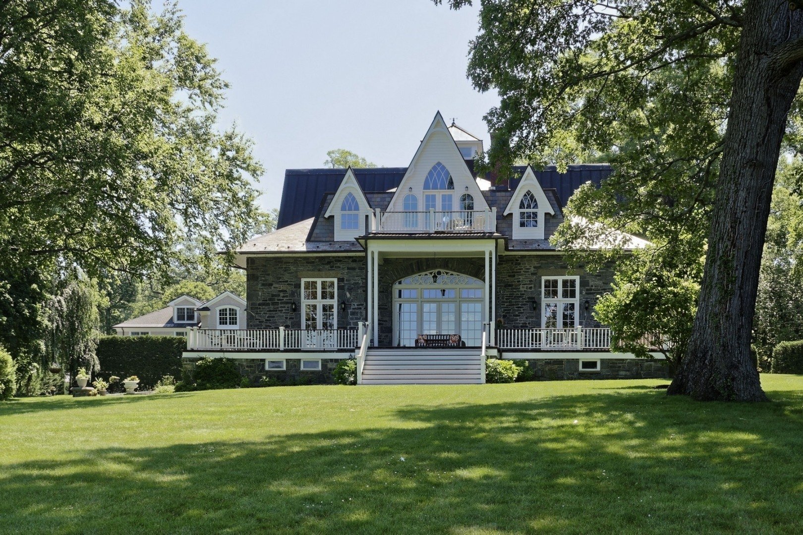 A large house with a dark stone exterior, multiple pointed windows, and a balcony on the upper level. It is surrounded by a well-maintained green lawn and tall trees, with sunlight filtering through the leaves.