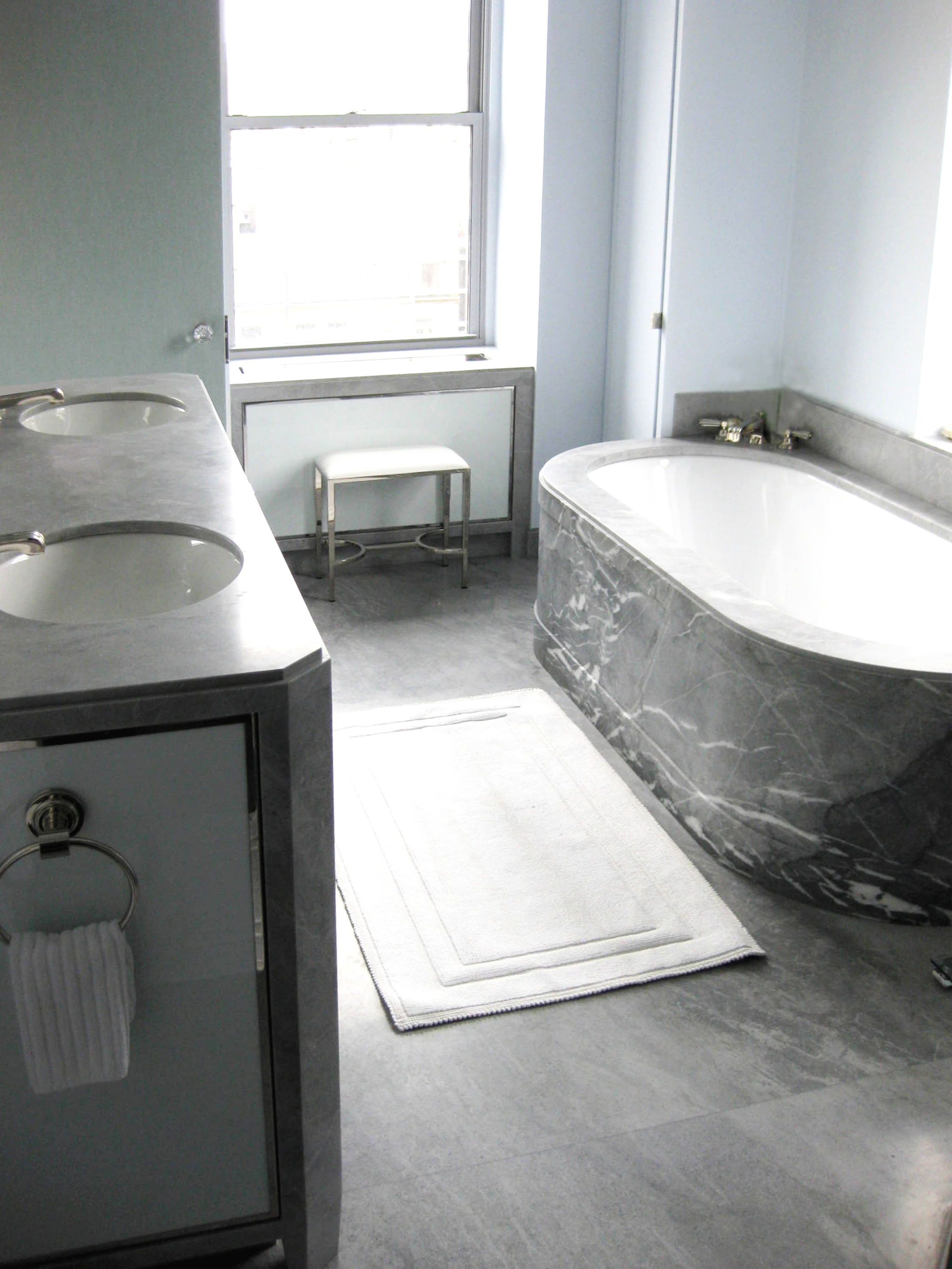 A bathroom with two sinks on the left, a window, a white stool, and a large marble bathtub on the right.