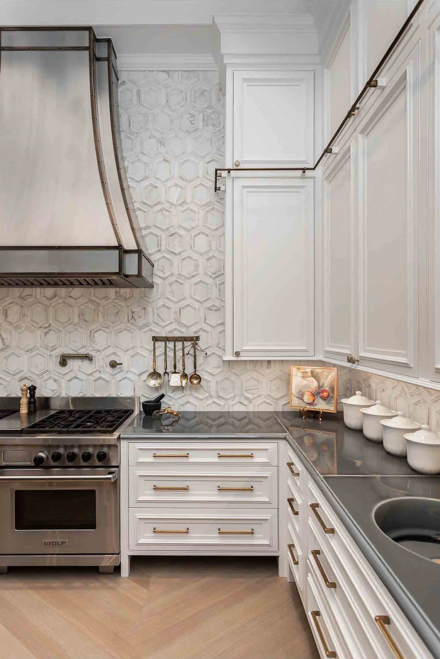 A corner of a modern kitchen with white cabinets, black countertops, and a stainless steel stove. The wall has textured tile with a honeycomb pattern, and there are hanging kitchen utensils on a small rail. The countertop has some decorative jars, a 