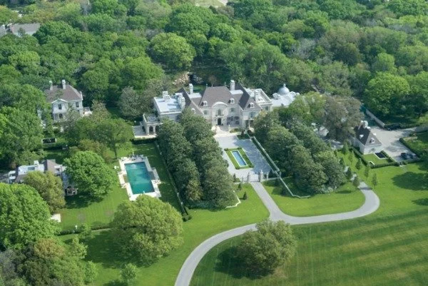 A large mansion surrounded by trees, with a swimming pool, a garden, and a driveway in a lush green landscape.