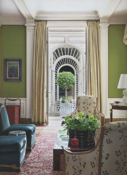 Interior of a living room with green walls, decorated with curtains, artwork, and antique furniture, featuring an arched doorway opening to an outdoor patio with a small tree and flowers.