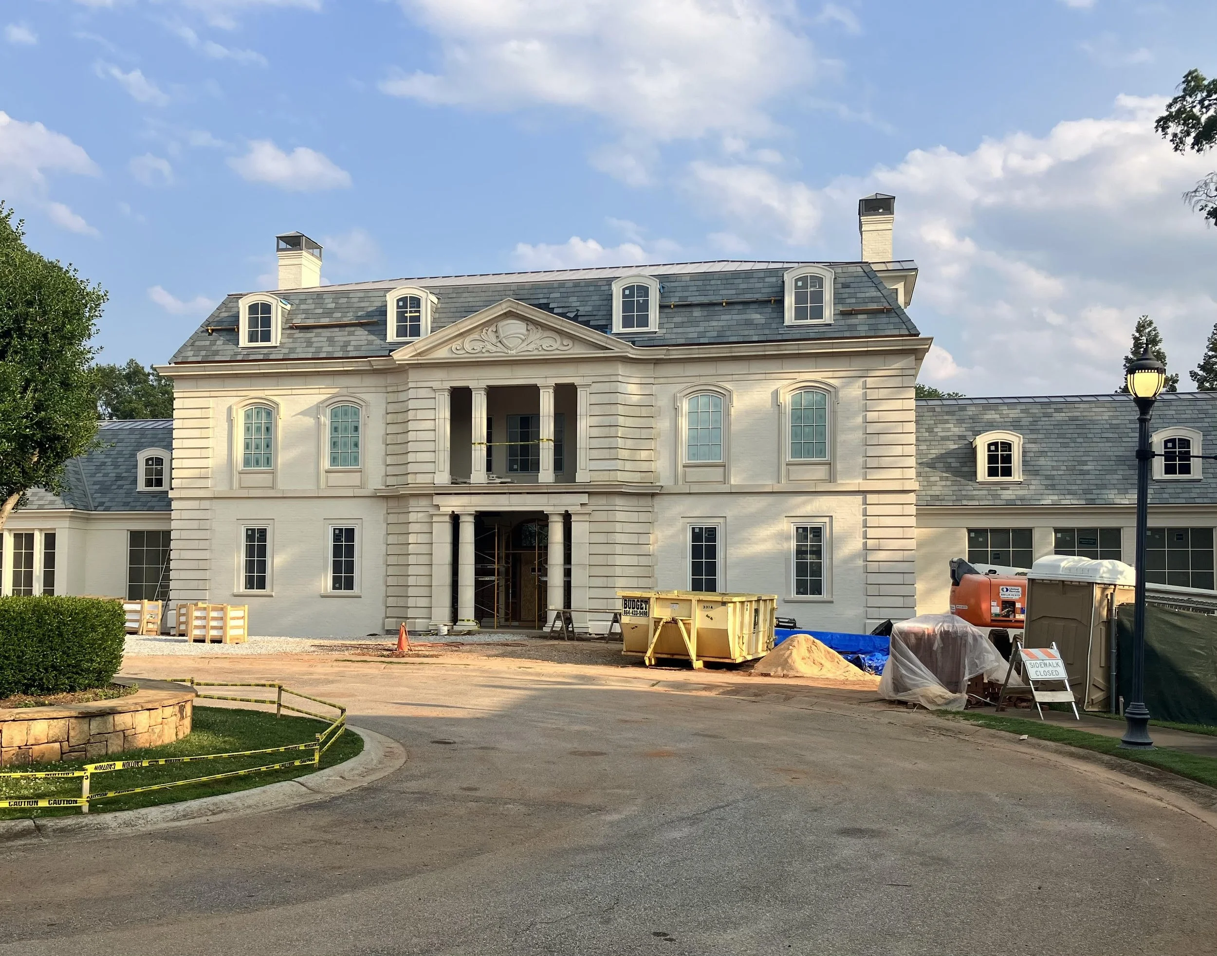 Under construction large white mansion with a classical design, multiple windows, and a prominent central porch area, surrounded by construction equipment and a caution tape barrier.