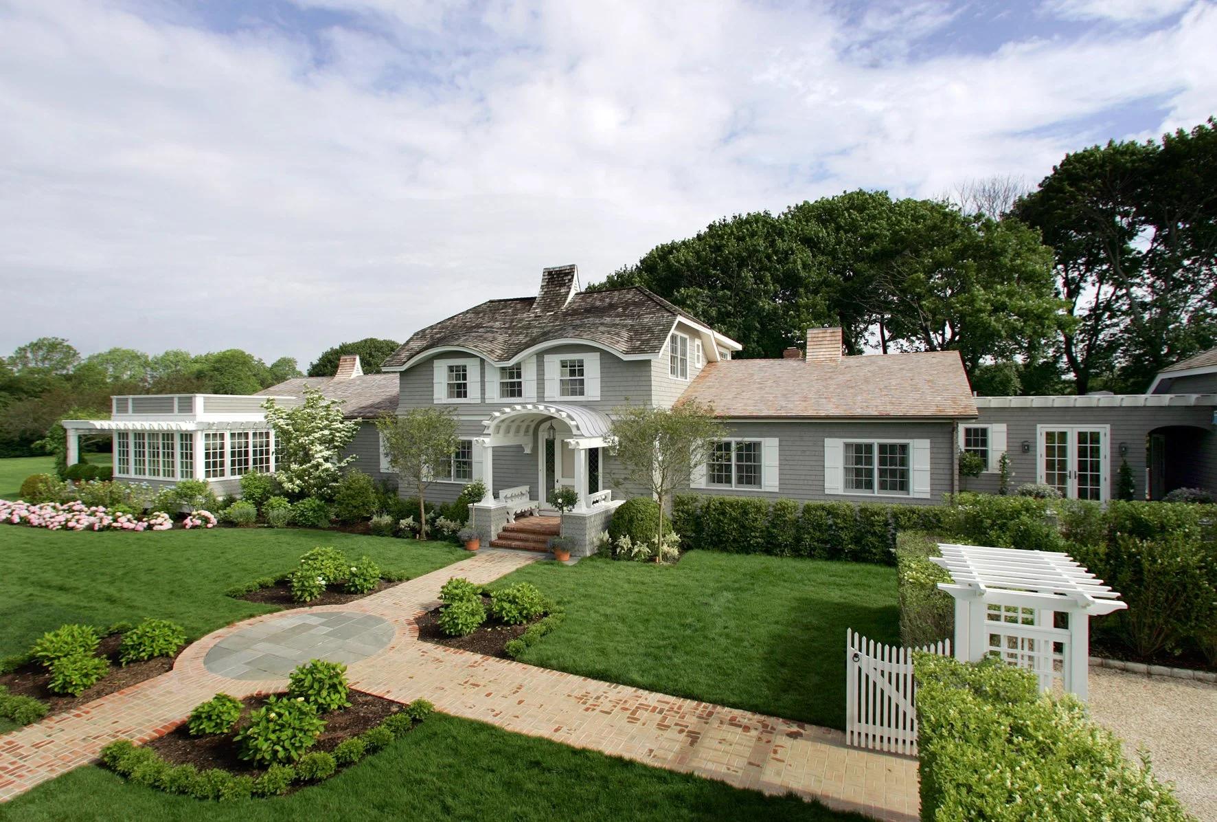 Large gray house with a curved roof, front porch, and multiple white-trimmed windows, surrounded by a well-maintained lawn, garden, and brick walkway under a partly cloudy sky.