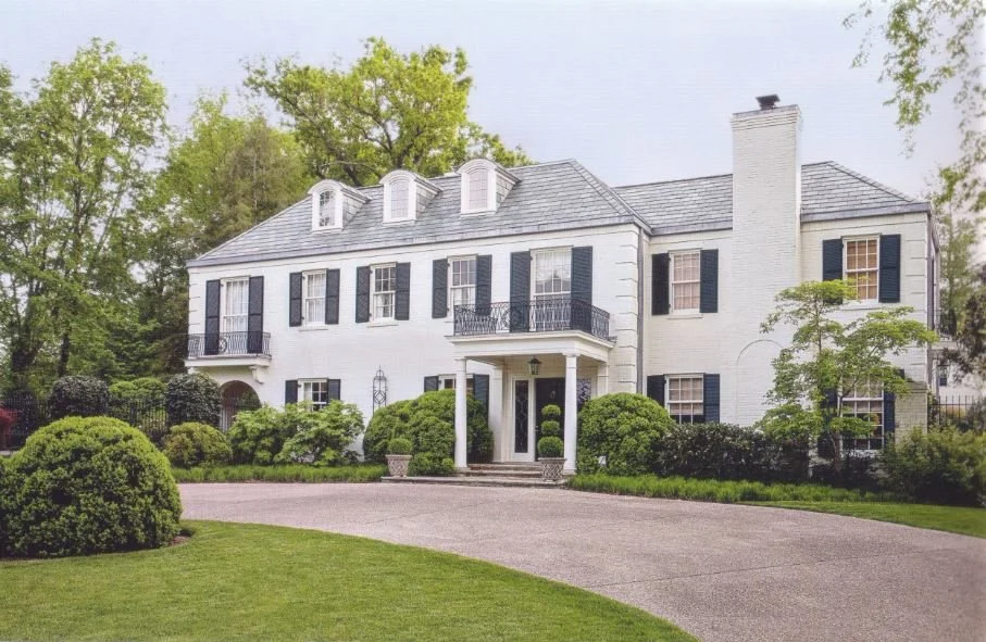 White two-story house with black shutters, front porch, and a driveway, surrounded by well-maintained lawn and trees.